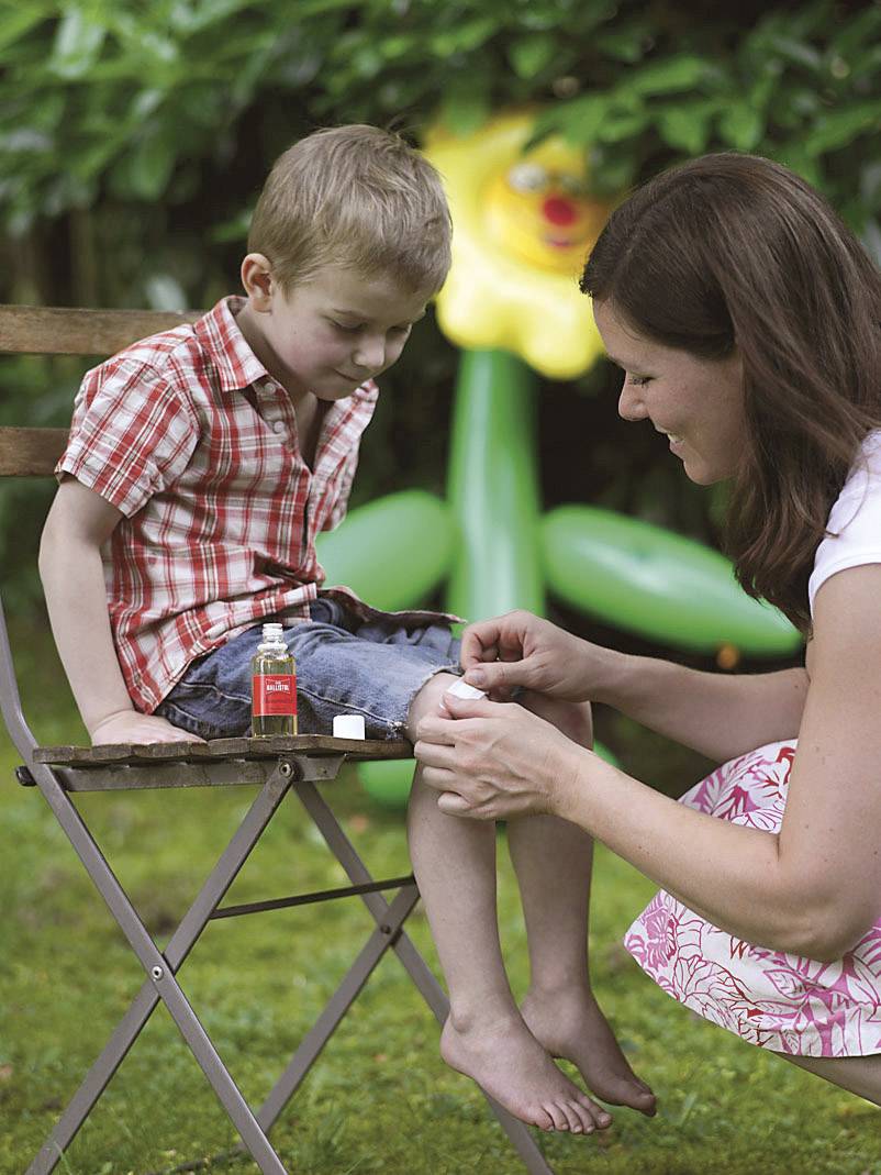 Eine Frau verarztet einen Jungen mit einem Pflaster auf dem Knie. Der Junge sitzt auf einem Stuhl im Garten.