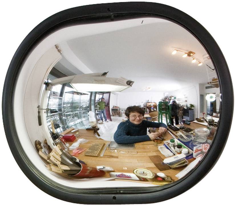 Eine Person sitzt lächelnd an einem Tisch in einem Raum voller technischer Geräte und Modelle, aufgenommen durch einen Fischaugen-Spiegel.