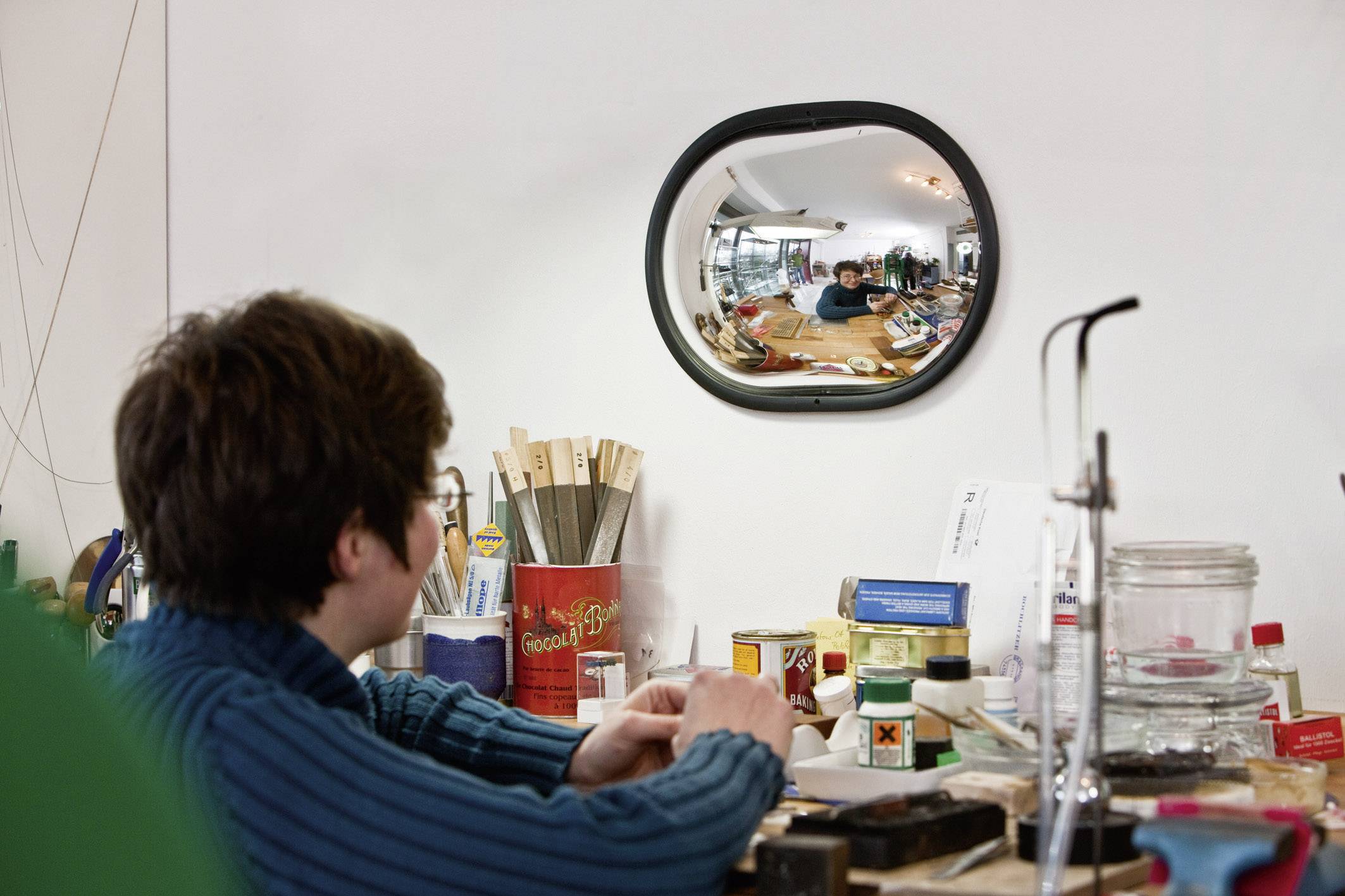 Eine Person sitzt an einem Tisch voller Materialien in einer Werkstatt und schaut in einen gewölbten Spiegel, der den Raum reflektiert.
