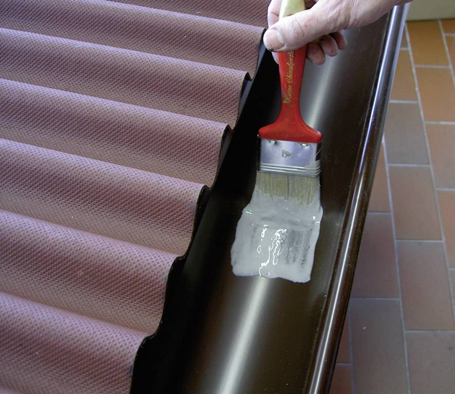 Eine Person trägt mit einem Pinsel eine transparente Flüssigkeit auf eine wellige Dachrinne auf.