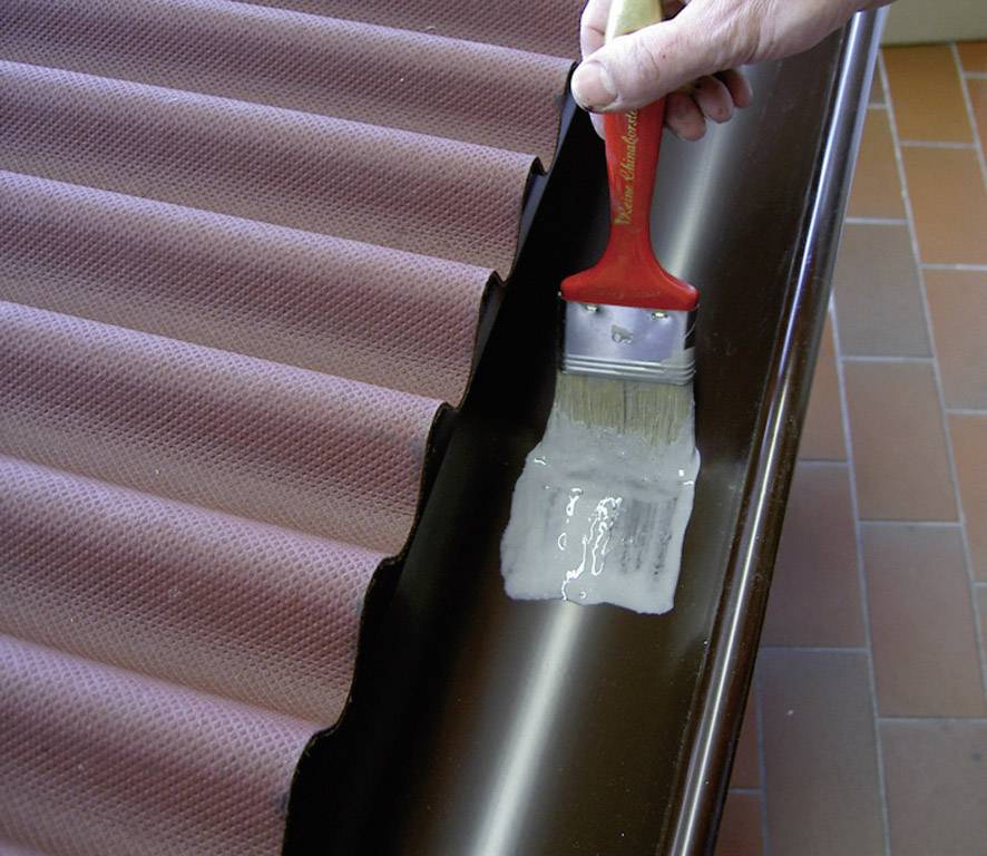 Eine Hand trägt mit einem Pinsel eine flüssige Beschichtung auf eine Dachrinne neben einem gewellten, rotbraunen Dach auf.
