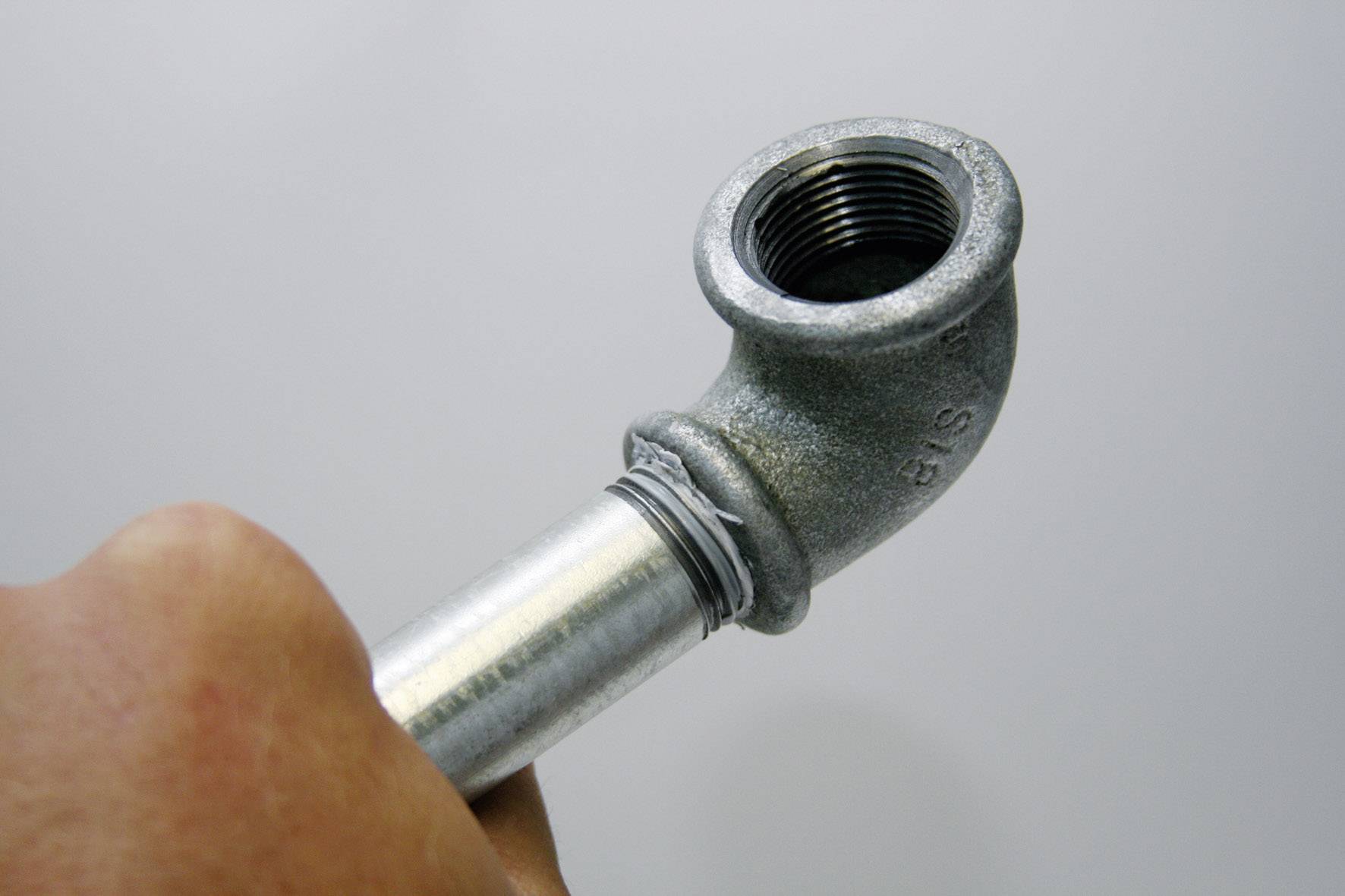 Eine Hand hält ein L-förmiges Metallrohr mit einem Gewinde. Das Rohr ist graumetallic und hat eine gebogene Form.