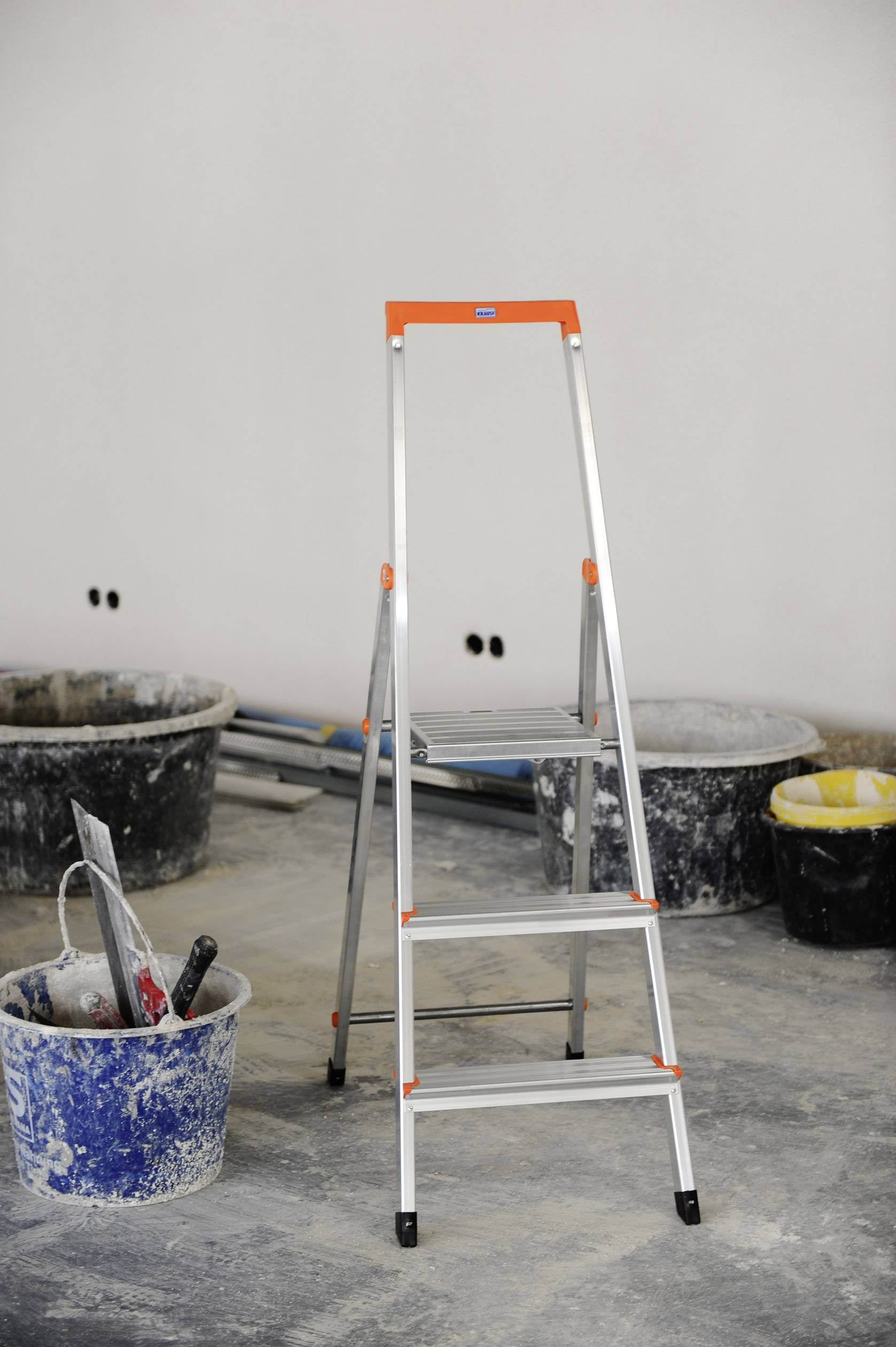 Leiter auf einer Baustelle, umgeben von Eimern und Werkzeugen für Malerarbeiten. Wand im Hintergrund ist noch unfertig.