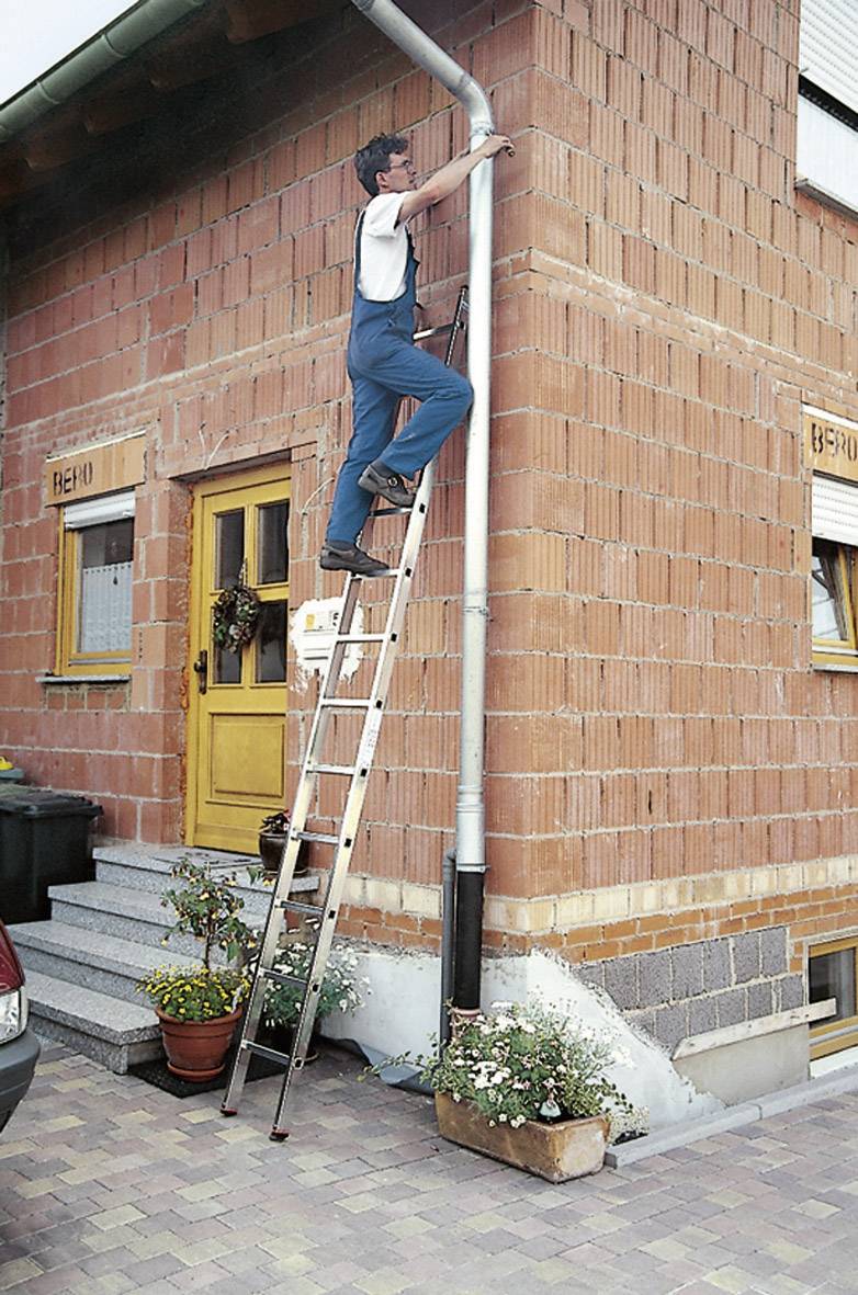 Ein Mann steht auf einer Leiter und repariert eine Dachrinne an einem Ziegelhaus. Ein Blumenkasten steht neben der Haustür.