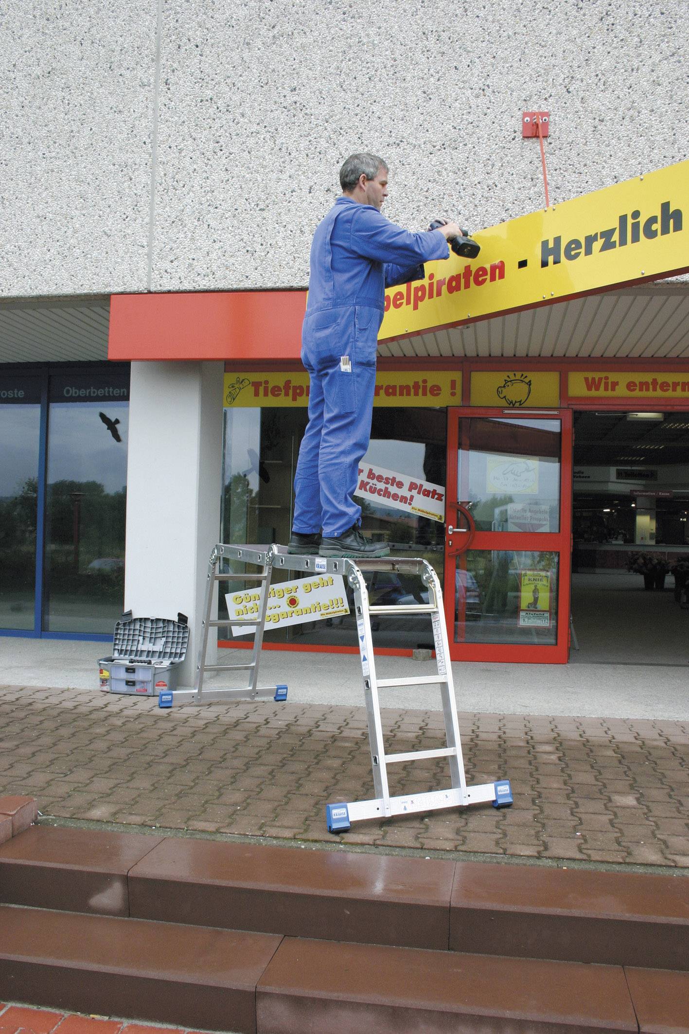 Ein Mann in blauer Arbeitskleidung steht auf einer klappbaren Leiter und arbeitet an einem Schild über dem Eingang eines Gebäudes.