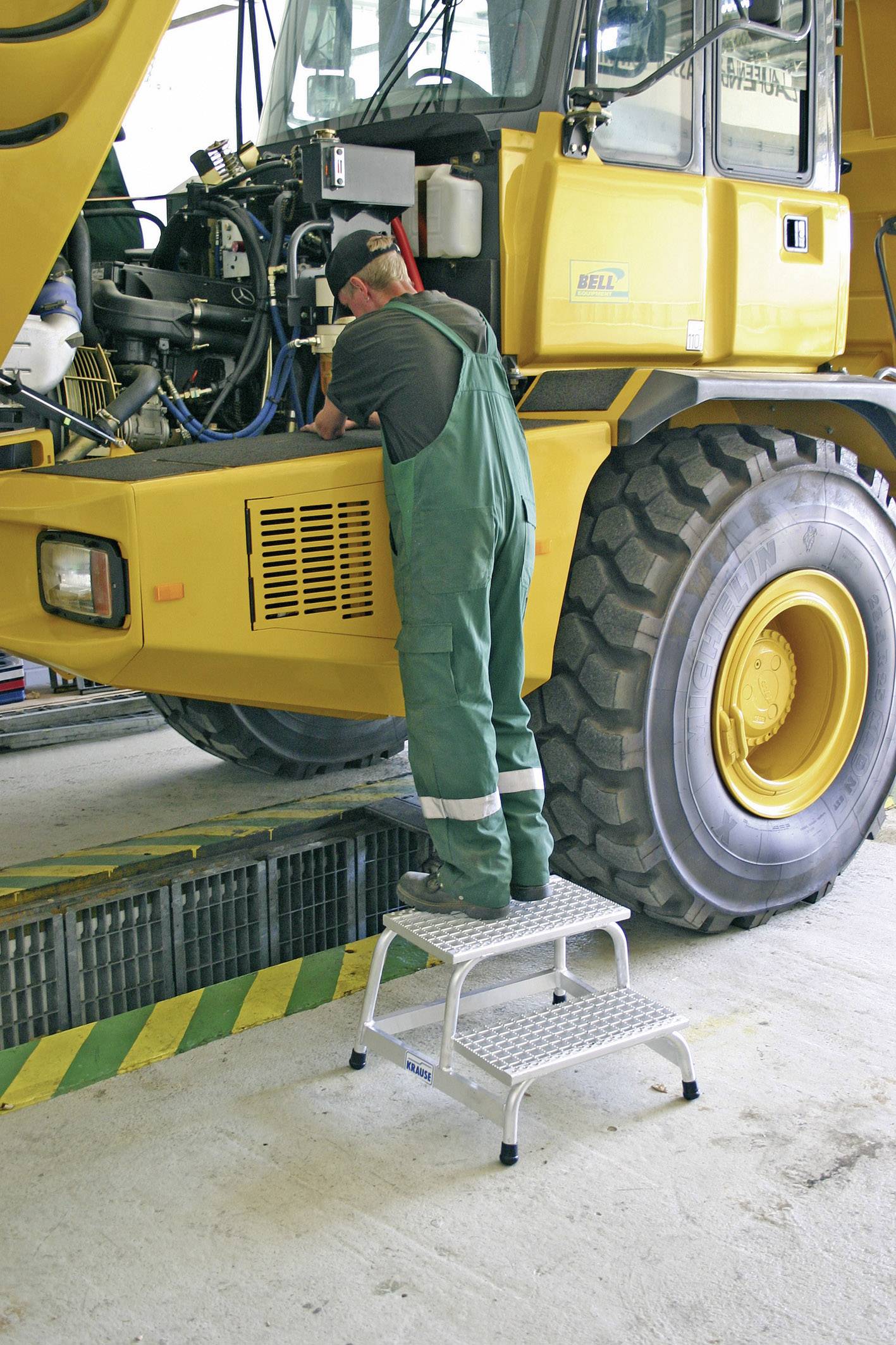 Ein Mechaniker in grüner Arbeitskleidung steht auf einer Trittleiter und repariert einen großen gelben Kran in einer Werkstatt.