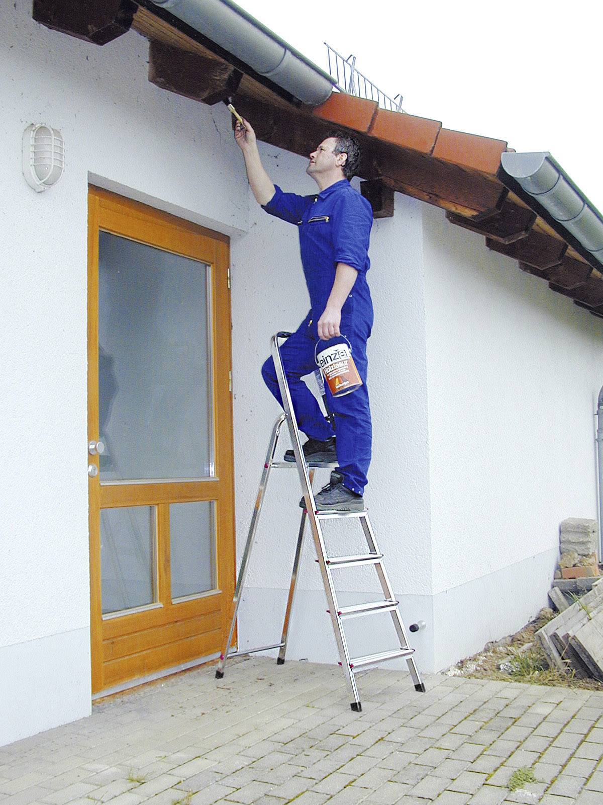 Mann in blauer Arbeitskleidung steht auf einer Leiter und streicht Holz am Dach eines Hauses. Neben ihm steht ein Eimer mit Farbe.