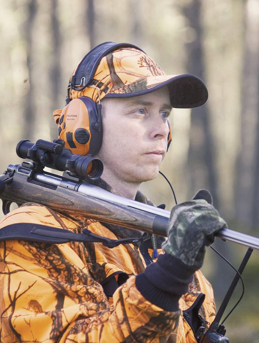 Eine Person in orangefarbener Jagdbekleidung trägt ein Gewehr und trägt Kopfhörer, während sie aufmerksam in einem bewaldeten Gebiet steht.