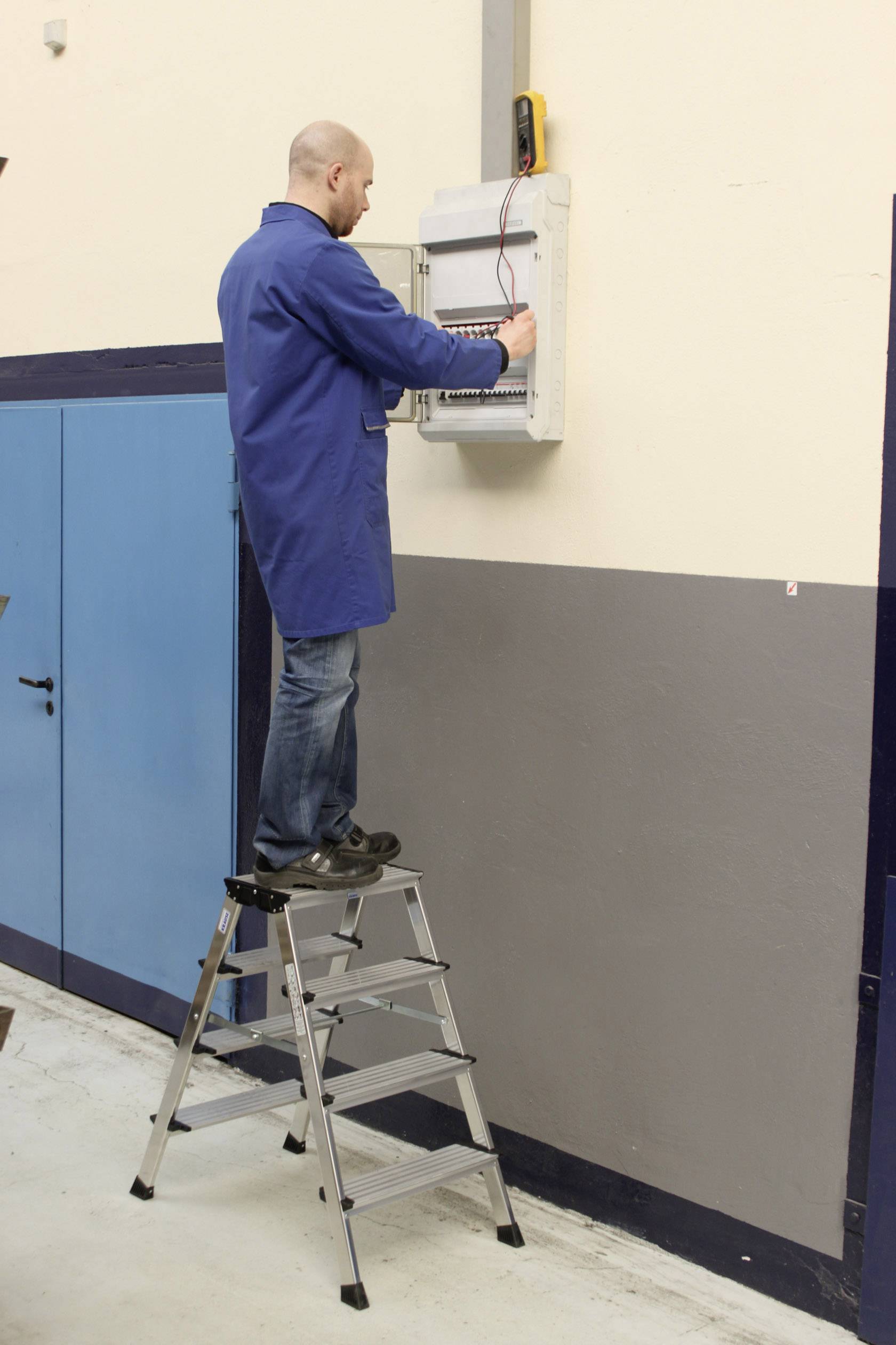 Ein Mann in einem blauen Kittel steht auf einer Leiter und arbeitet an einem elektrischen Sicherungskasten an der Wand.