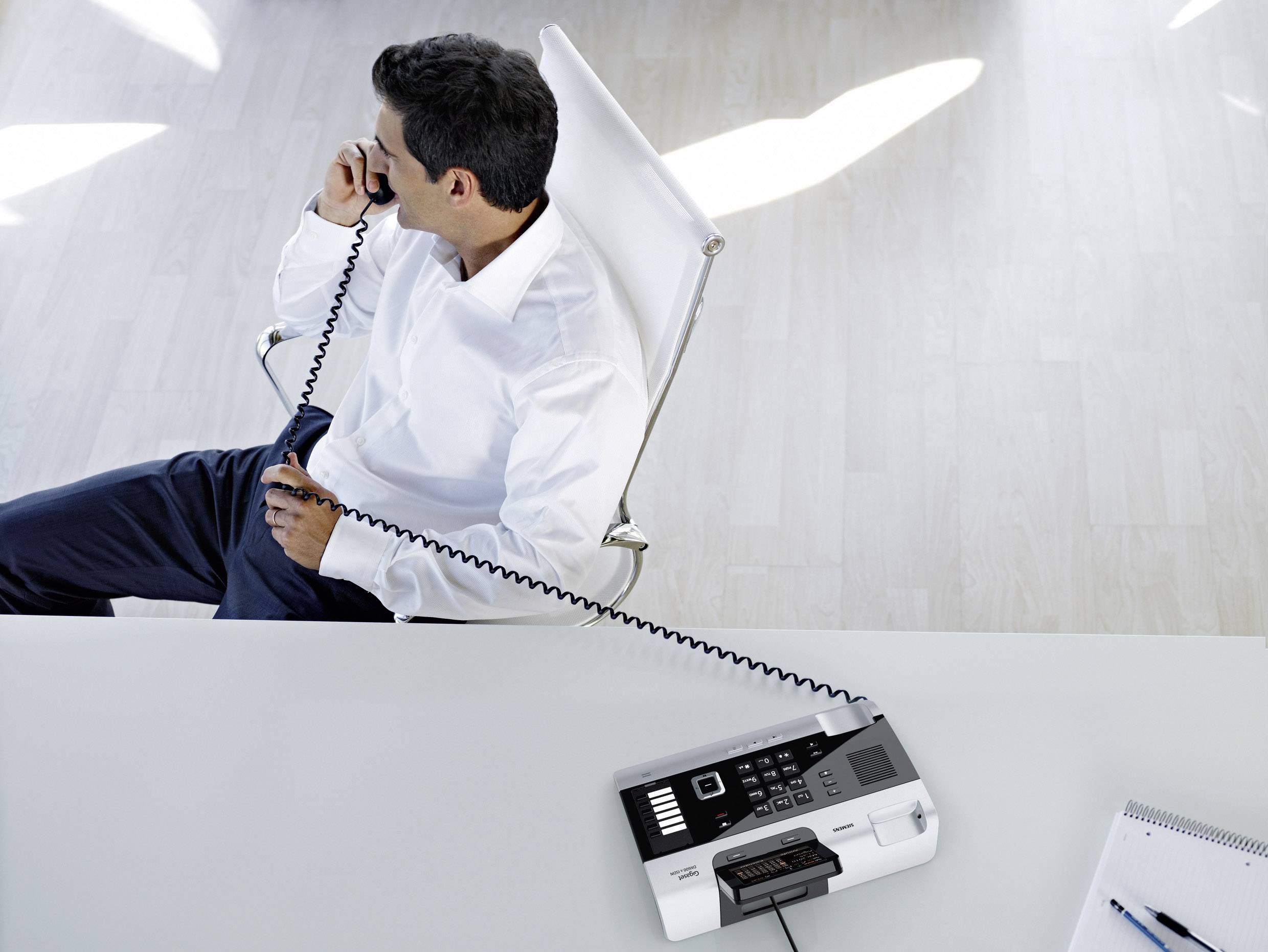 Ein Mann sitzt auf einem Stuhl und spricht in ein Telefon. Auf dem Tisch steht ein Faxgerät. Im Vordergrund liegt ein Notizbuch.