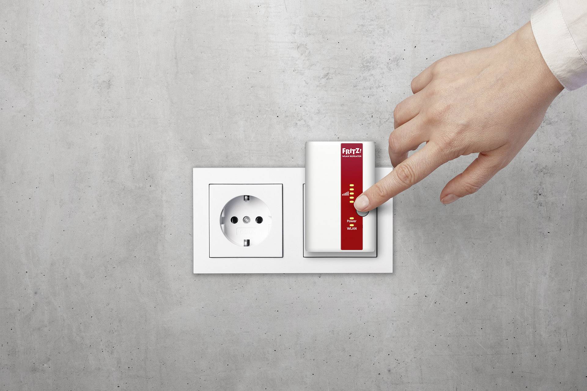Hand drückt auf den Knopf eines WLAN-Repeaters, der neben einer Steckdose an einer grauen Wand installiert ist.