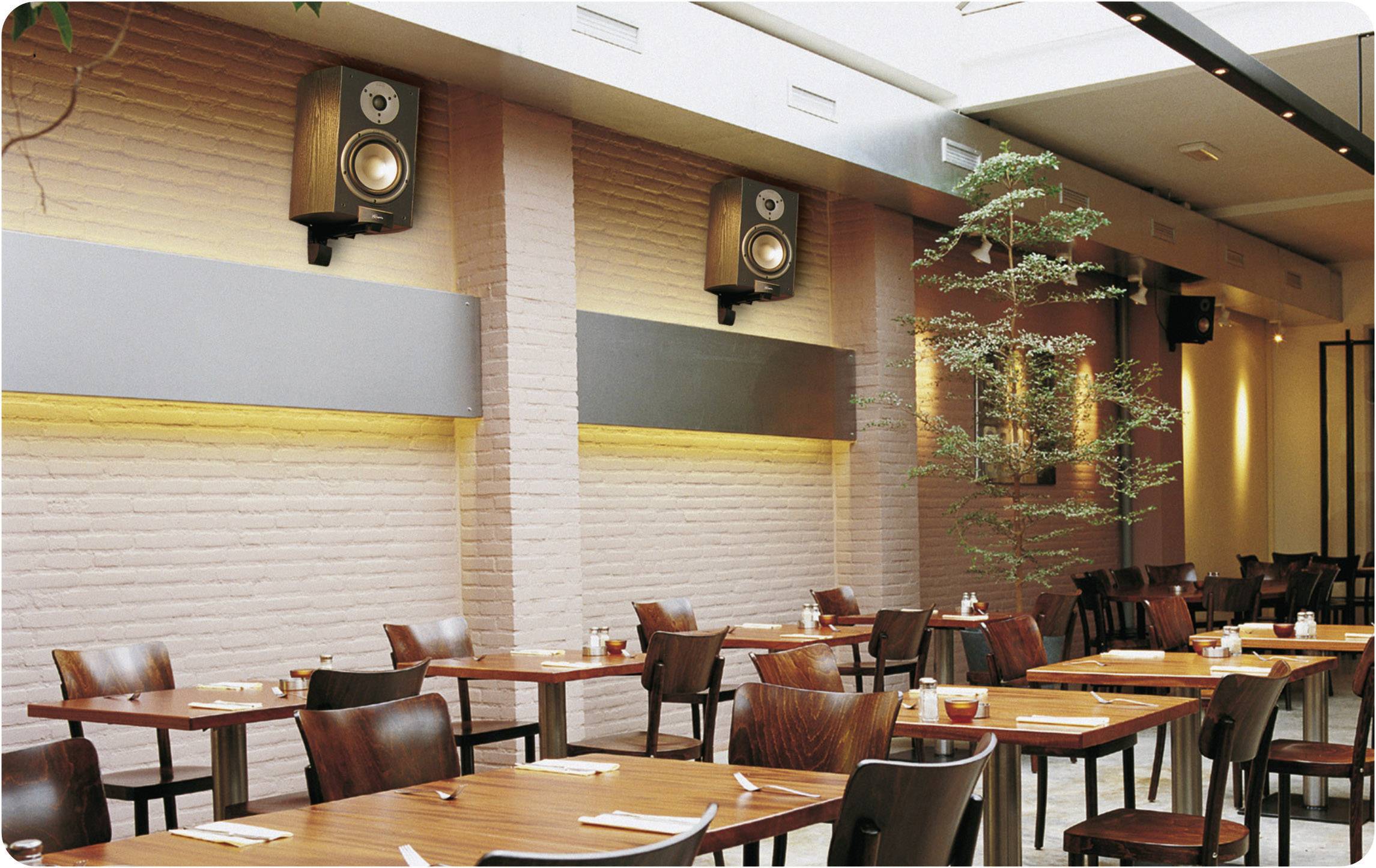 Ein modernes Restaurant mit Holztischen und -stühlen. An den Wänden befinden sich Lautsprecher. Große Fenster lassen viel Licht herein.