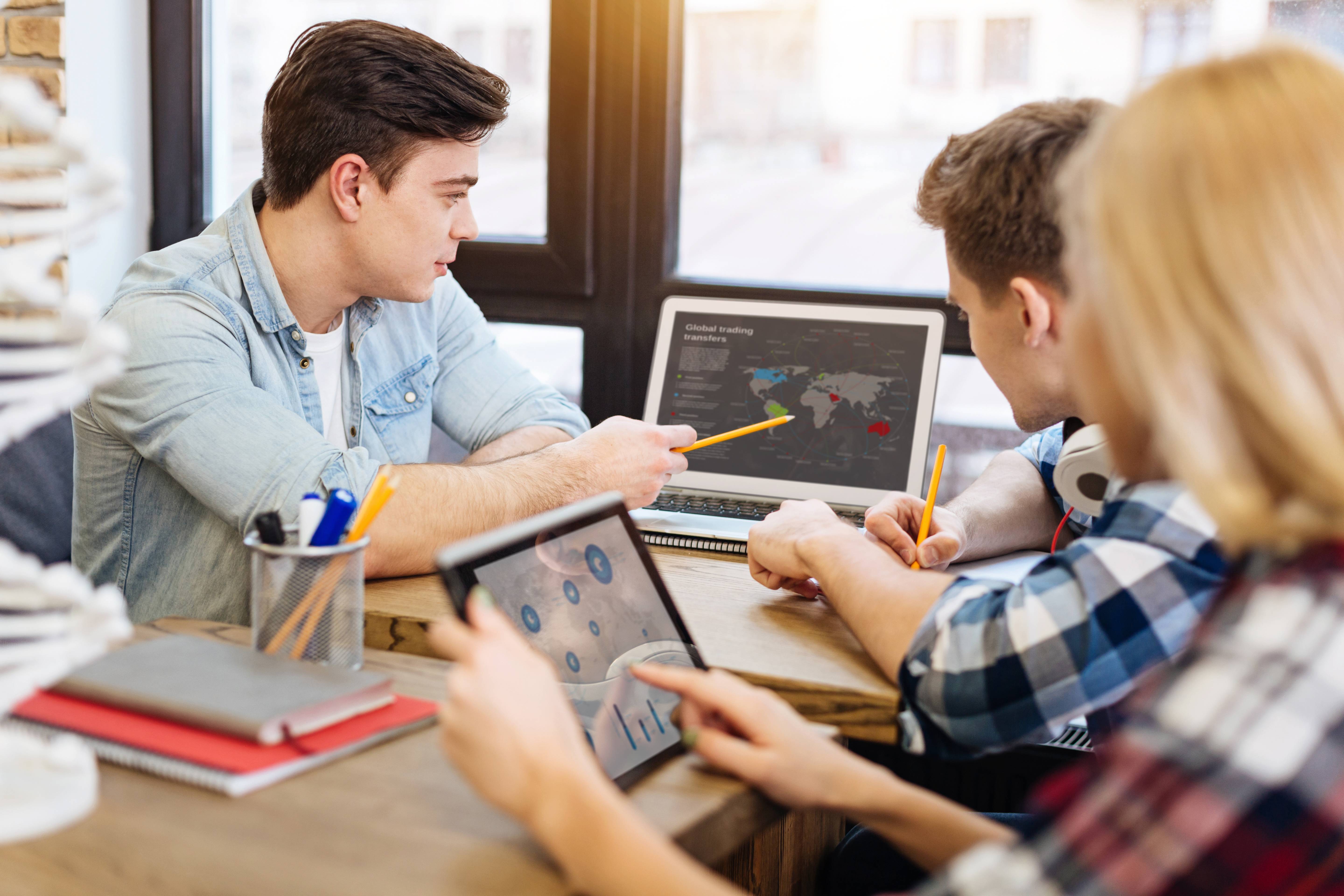 Drei Schüler arbeiten zusammen an einem Laptop und Tablet und besprechen Daten auf einer Weltkarte.