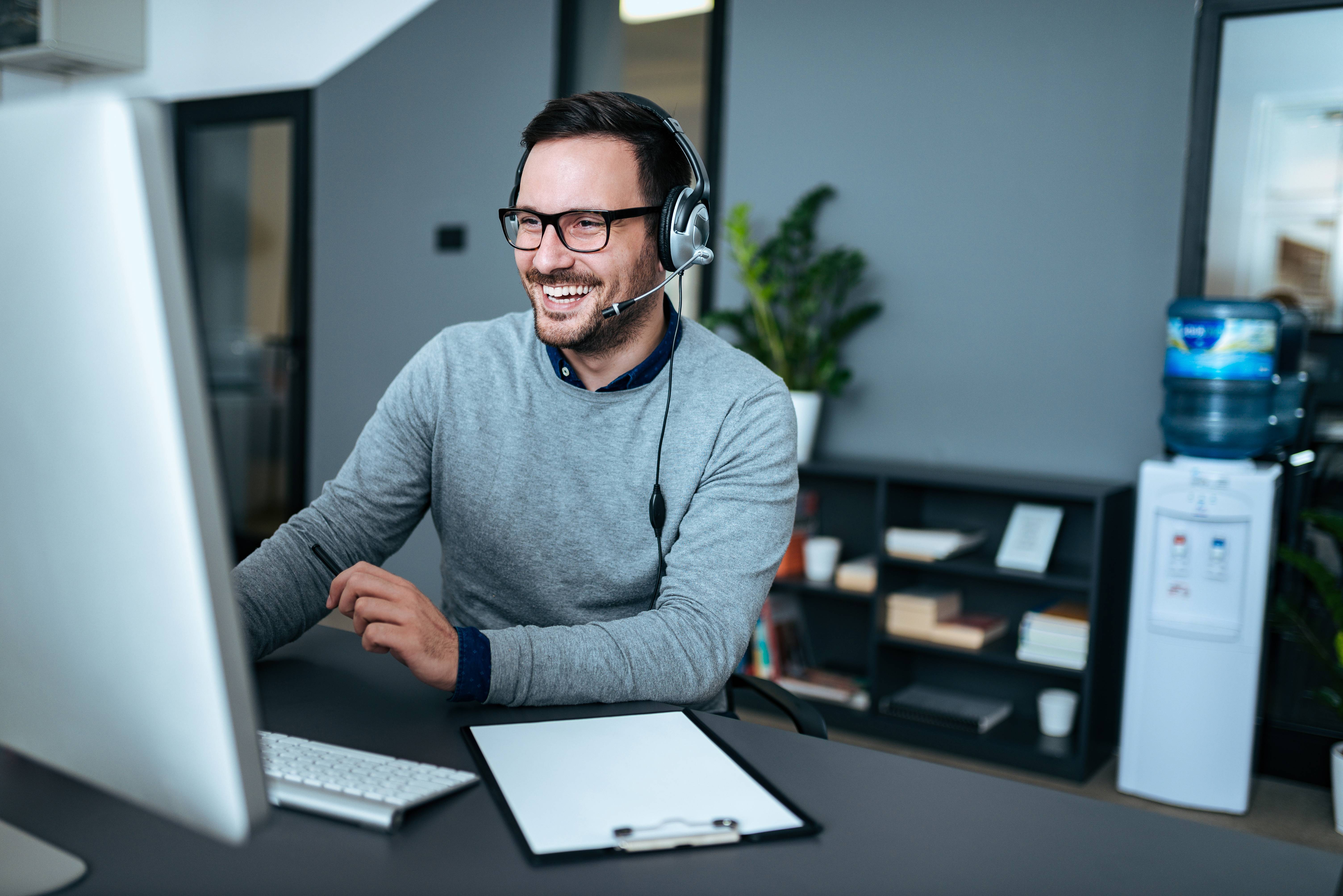 Mann mit Headset sitzt lächelnd am Schreibtisch und arbeitet am Computer.