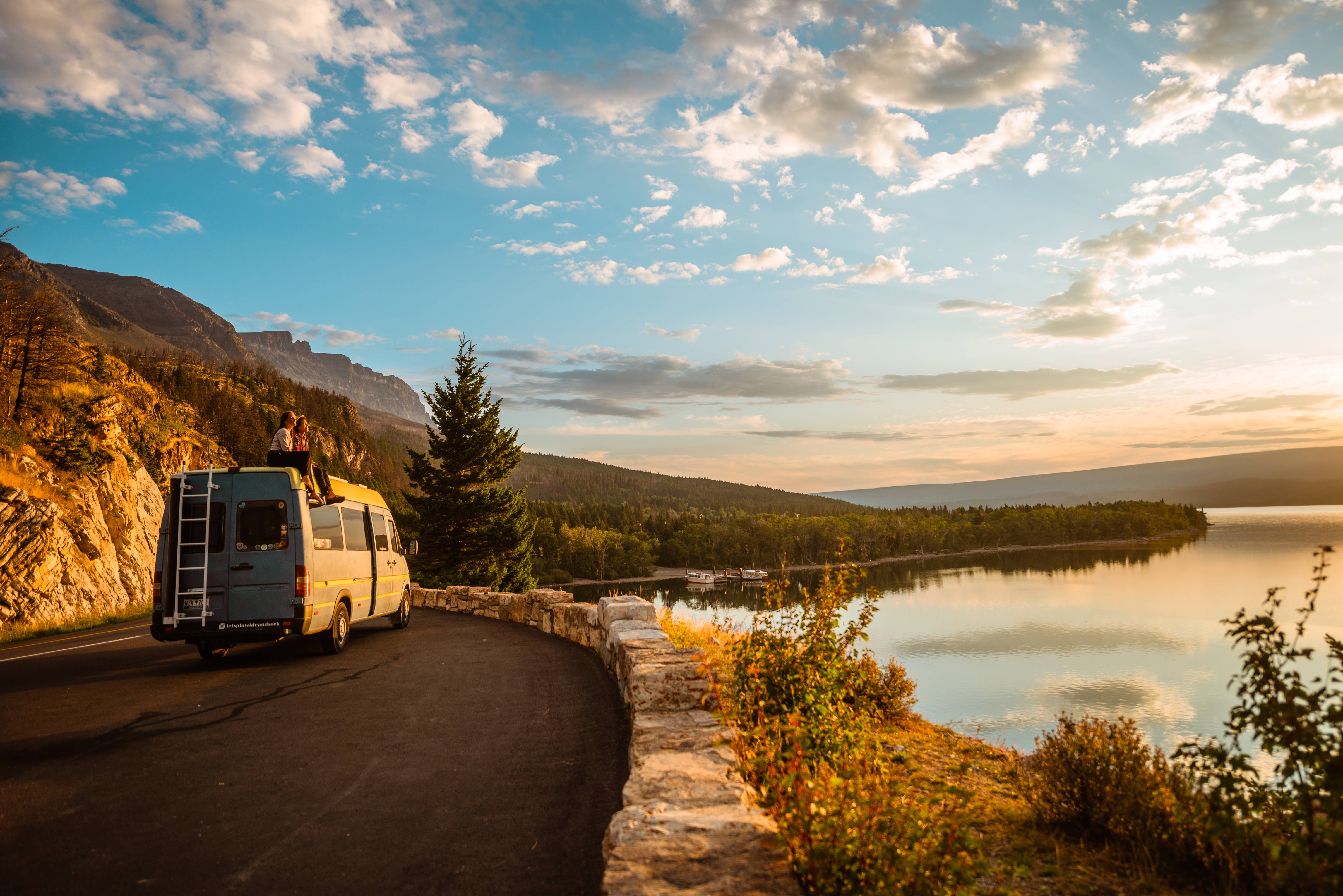 Ein Campervan steht auf einer Küstenstrasse bei Sonnenuntergang, zwei Personen sitzen auf dem Dach.