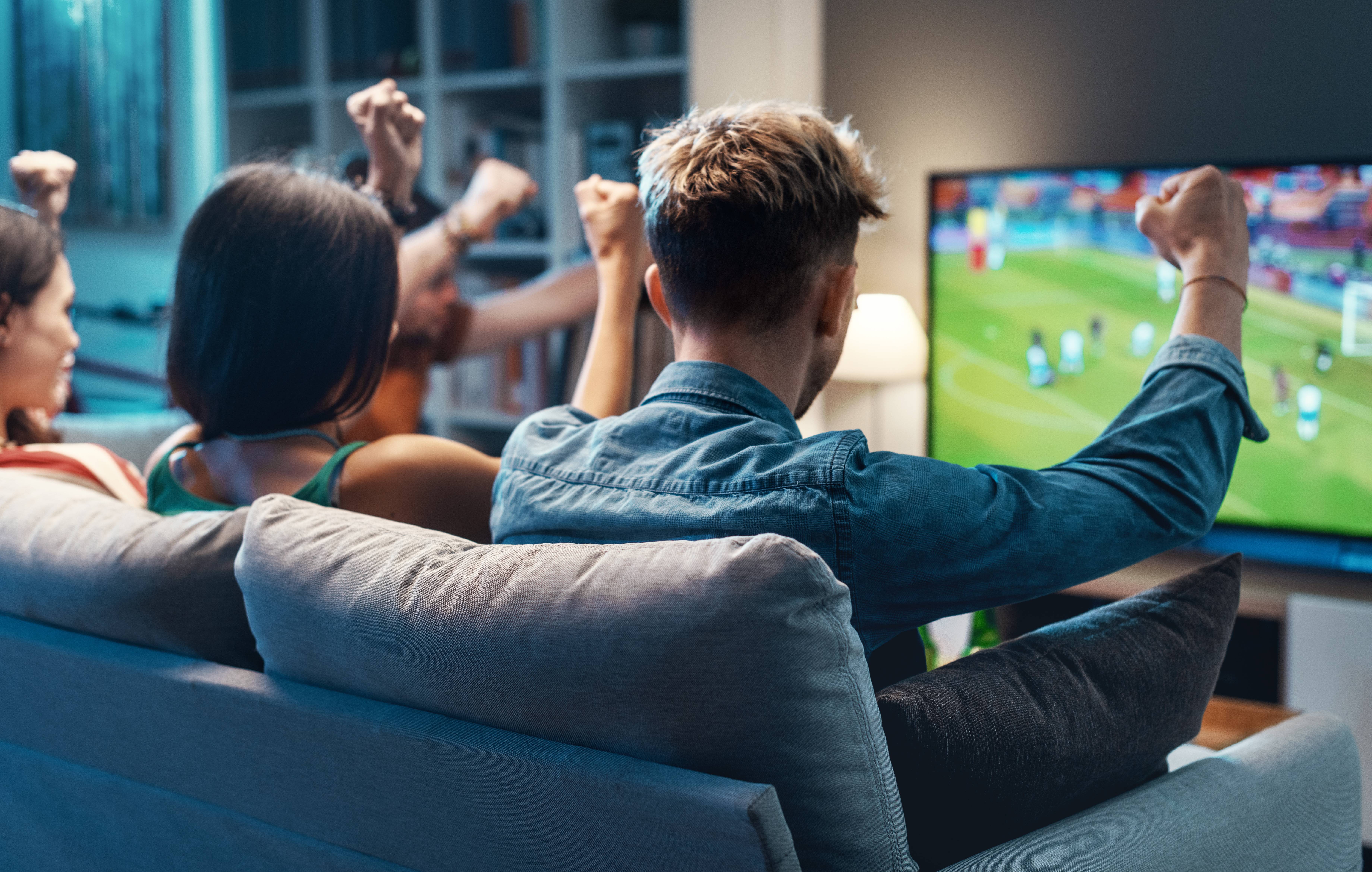 Freunde jubeln begeistert beim Ansehen eines Fußballspiels auf einem großen Fernseher im Wohnzimmer.