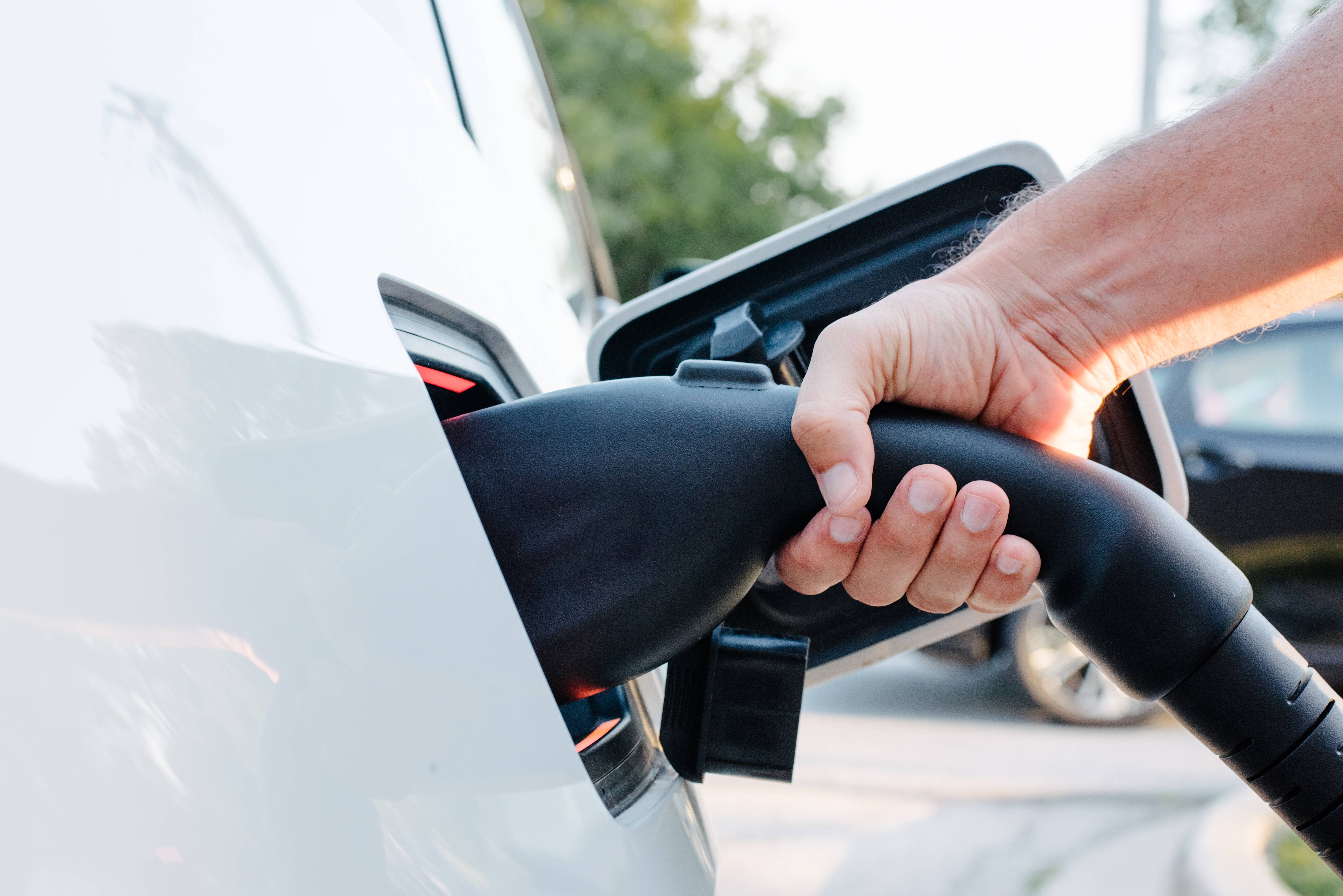 Nahaufnahme einer Hand, die den Ladestecker in die geöffnete Ladebuchse eines weißen E-Autos (EV) steckt. 
