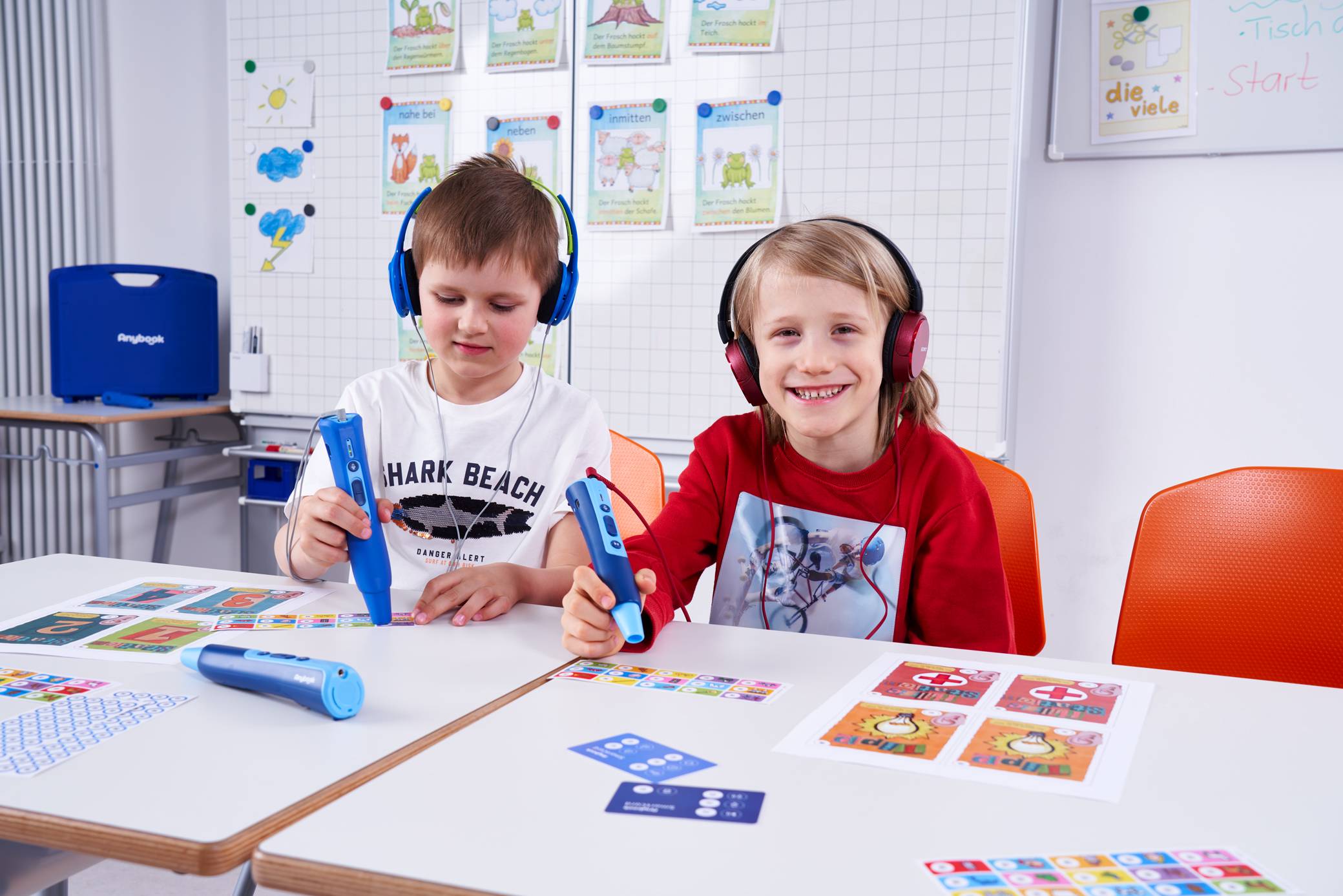 Zwei Kinder sitzen an einem Tisch und arbeiten mit dem Anybook Pro sowie dem Anybook Reader. Sie haben Kopfhörer auf.
