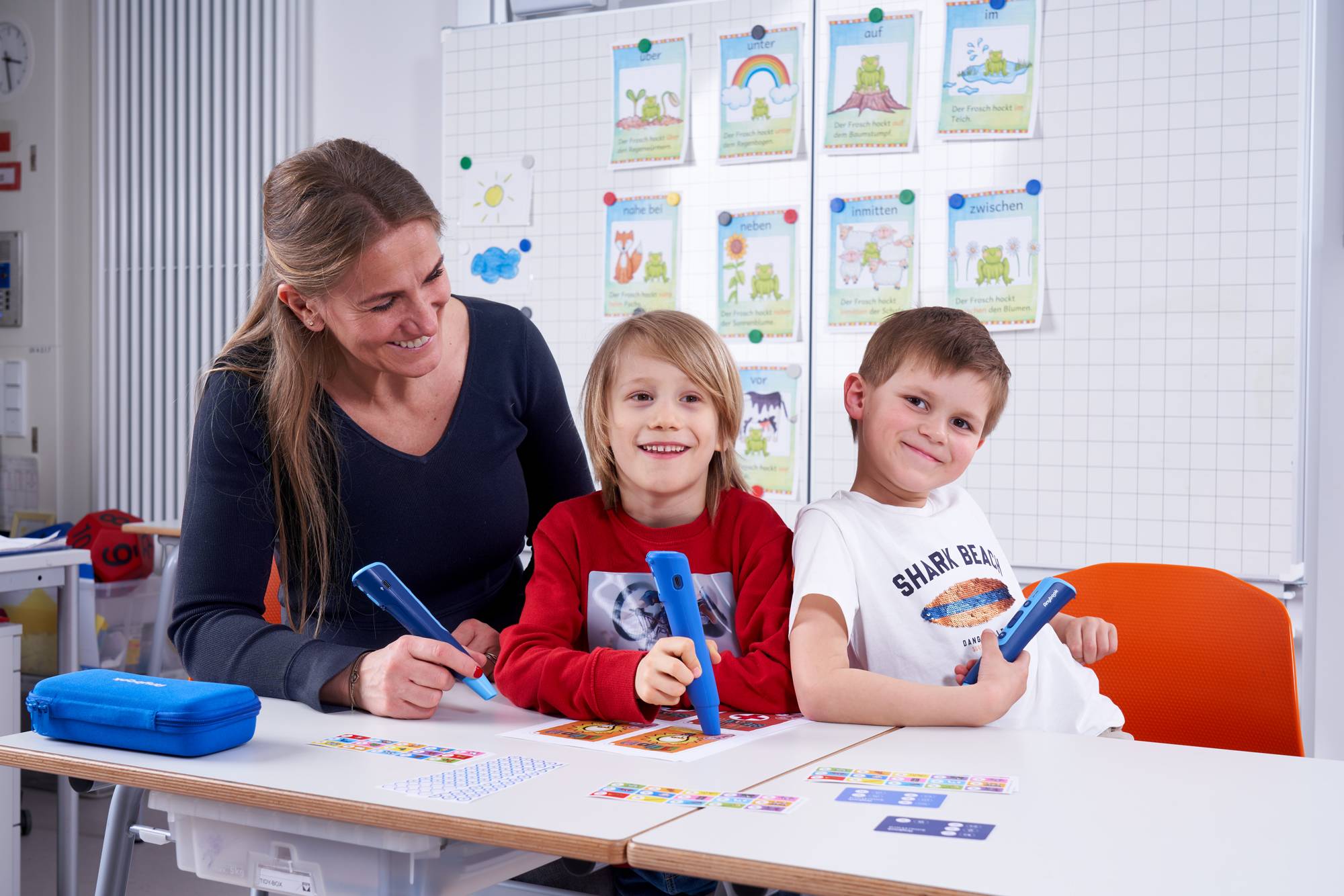 Zwei Kinder sitzen an einem Tisch und arbeiten mit dem Anybook Pro sowie dem Anybook Reader. Sie haben Kopfhörer auf.