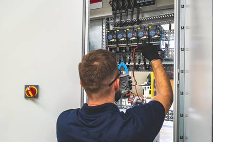 Das Bild zeigt eine Person, welche ein dunkelblaues Hemd, eine Schutzbrille und schwarze Handschuhe trägt und vor einem geöffneten Schaltschrank steht. Sie hält ein Multimeter mit roten und schwarzen Kabeln, die mit Komponenten im Inneren des Schaltschranks verbunden sind. Der Schrank enthält elektrische Bauteile, darunter Relais, Klemmenblöcke und Verkabelungen. Am Gehäuse des Schaltschranks ist ein rot-gelber Drehschalter verbaut.