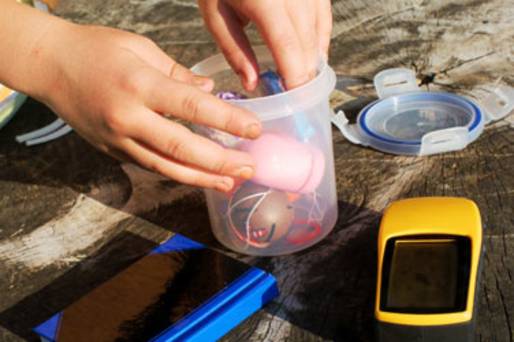 Ein Geocache, bestehend aus einer Dose, in der verschiedene Tauschgegenstände enthalten sind. Die Dose ist geöffnet und man sieht die Hände einer Person, die entweder gerade einen Gegenstand entnehmen oder hineinlegen möchte. Neben dem Behälter liegen ein zugeklapptes Logbuch und ein GPS-Gerät sowie der Dosendeckel. Der Untergrund besteht aus Holz. Es könnte sich dabei um einen Tisch handeln.