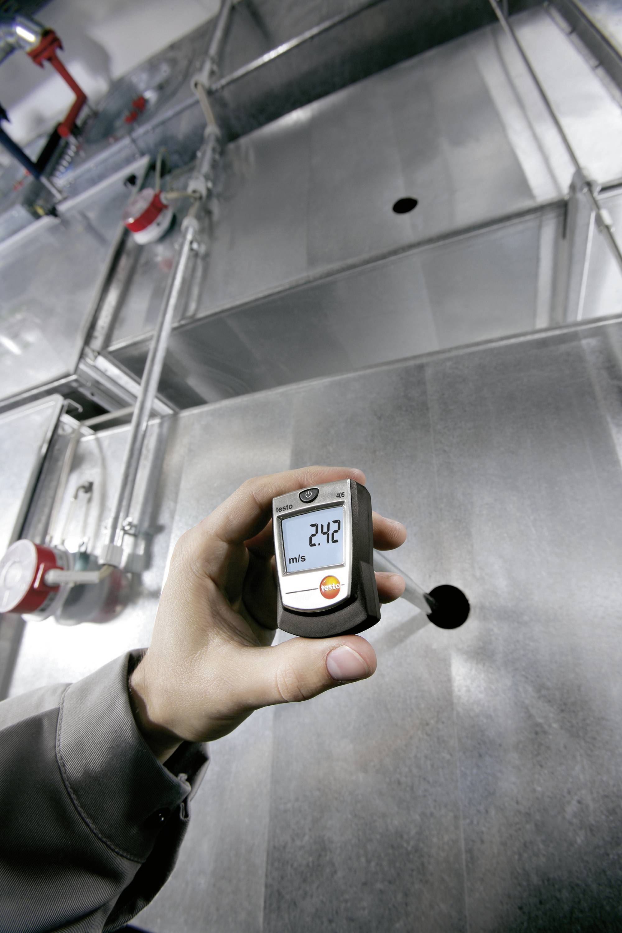A hand is holding a digital measuring device displaying '24.2'. Industrial ventilation pipes are visible in the background.