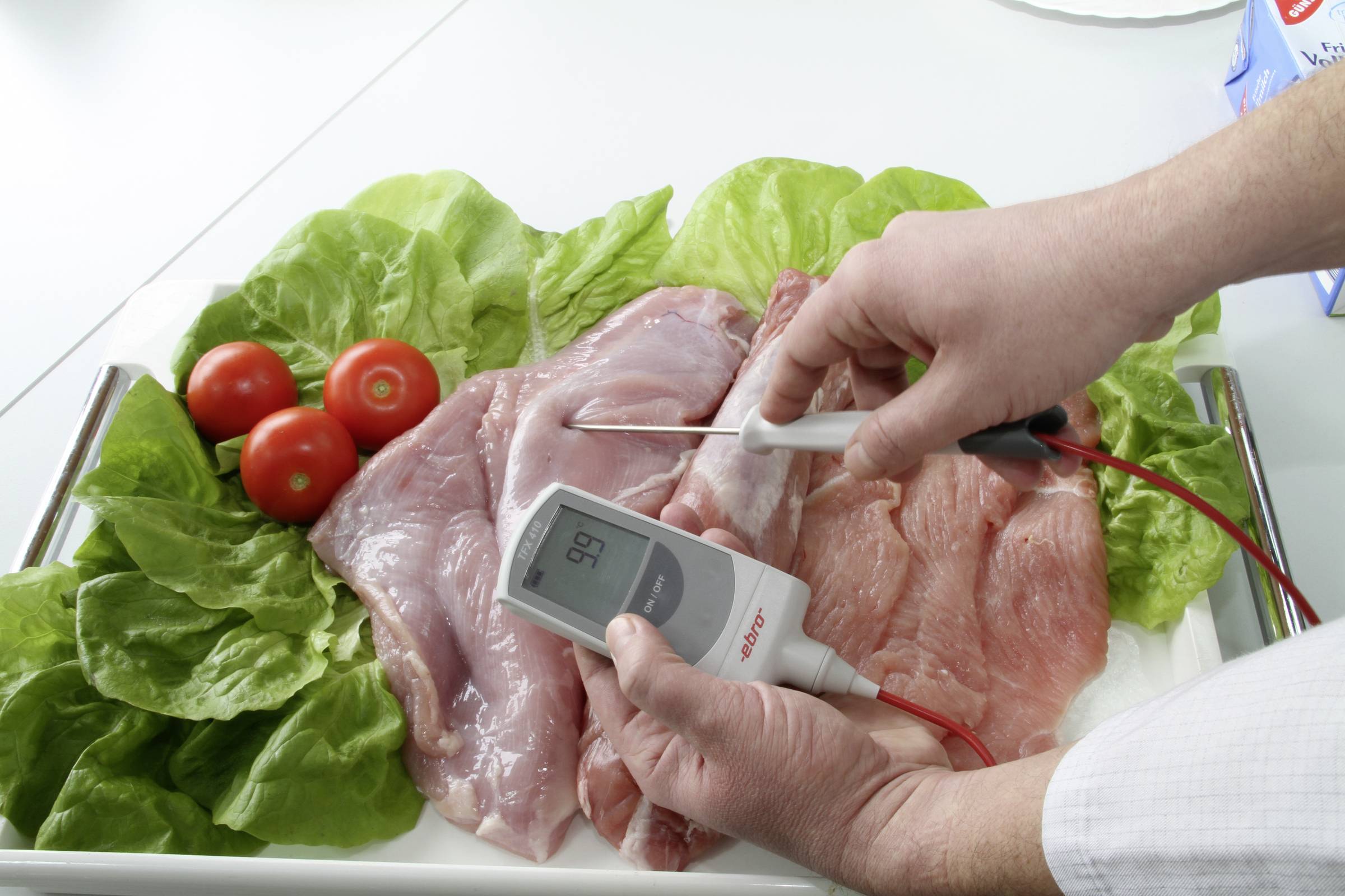 A person is measuring the temperature of raw meat on a plate with salad and tomatoes using a thermometer.