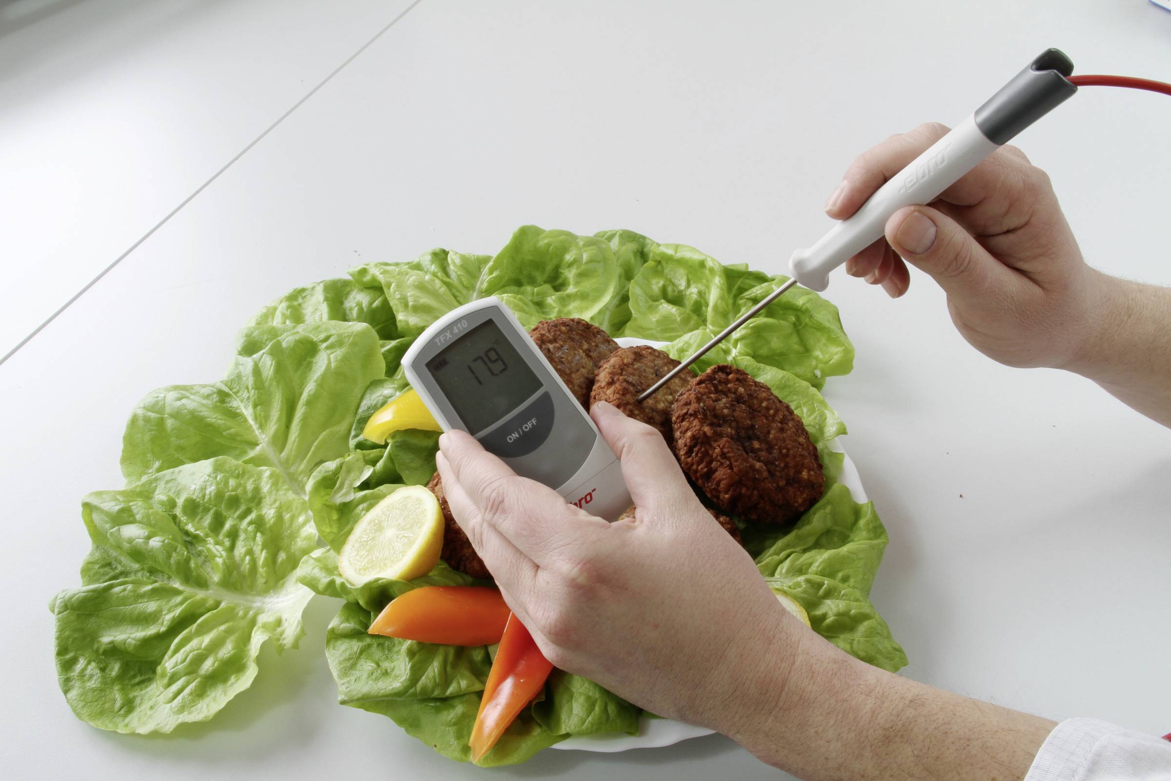 A person is measuring the temperature of meatballs on a lettuce leaf with pepper and lemon on a table using a thermometer.