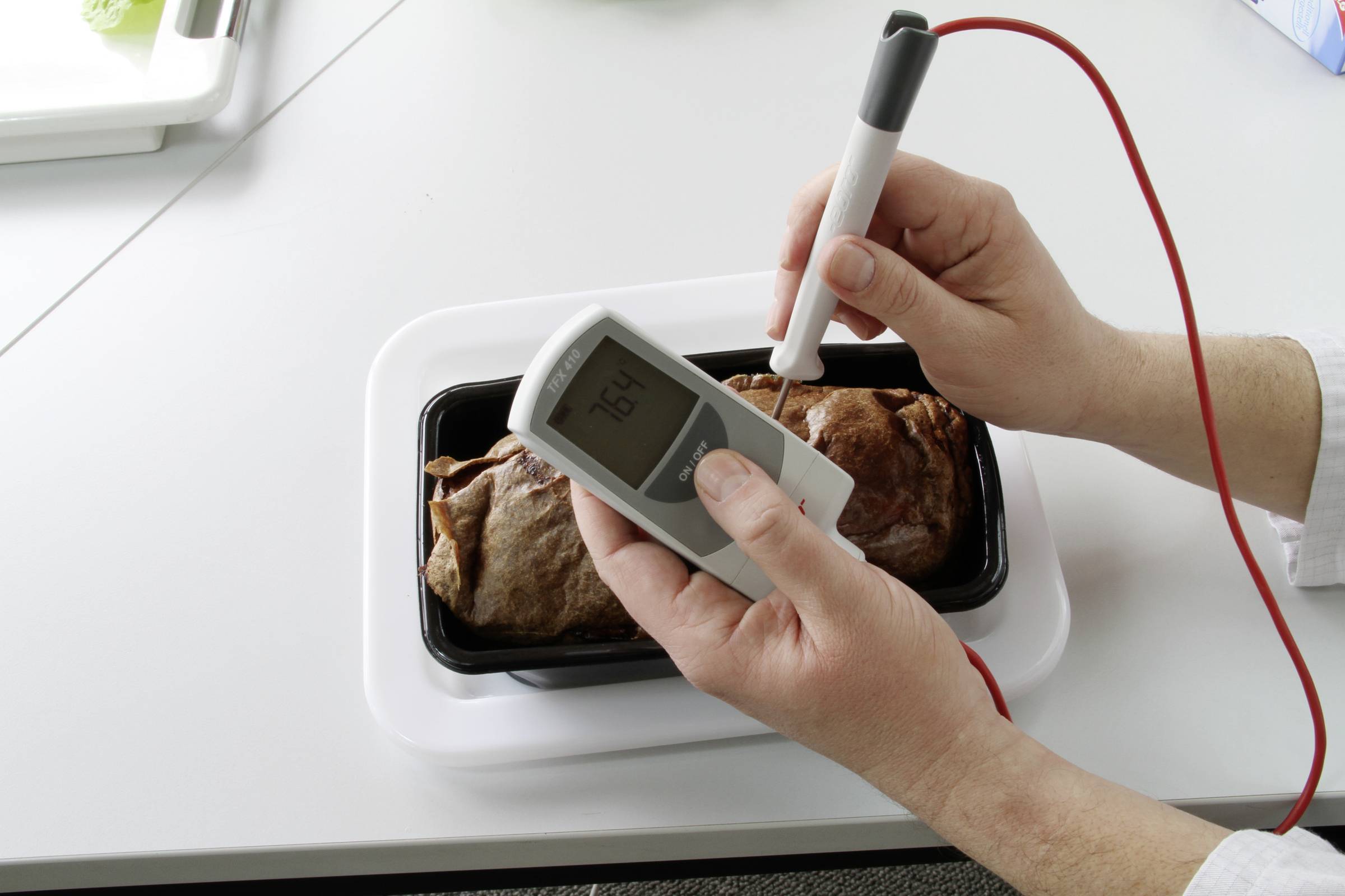 A person is measuring the core temperature of a roast in a black dish using a digital thermometer.