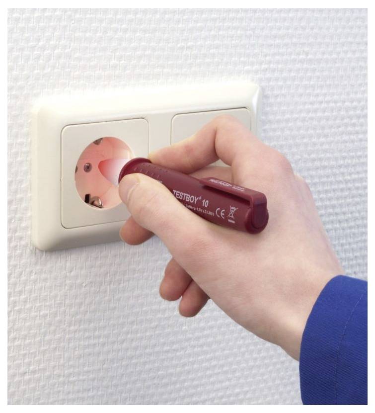 A hand holds a red voltage tester against an electrical outlet. The tester emits a red light, indicating the presence of voltage.