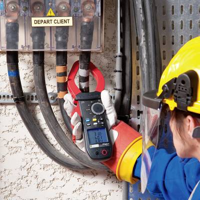A person in work attire is measuring electrical current at a control cabinet using a multimetre. Safety equipment is being worn.