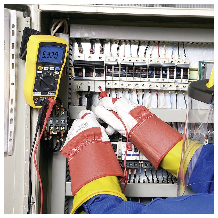 An electrician wearing protective clothing uses a multimeter to measure voltage in an open electrical cabinet.