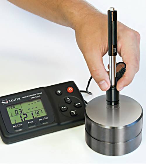 A hand holds a hardness testing device resting on a cylindrical metal block. A digital display device is positioned to the right of it.