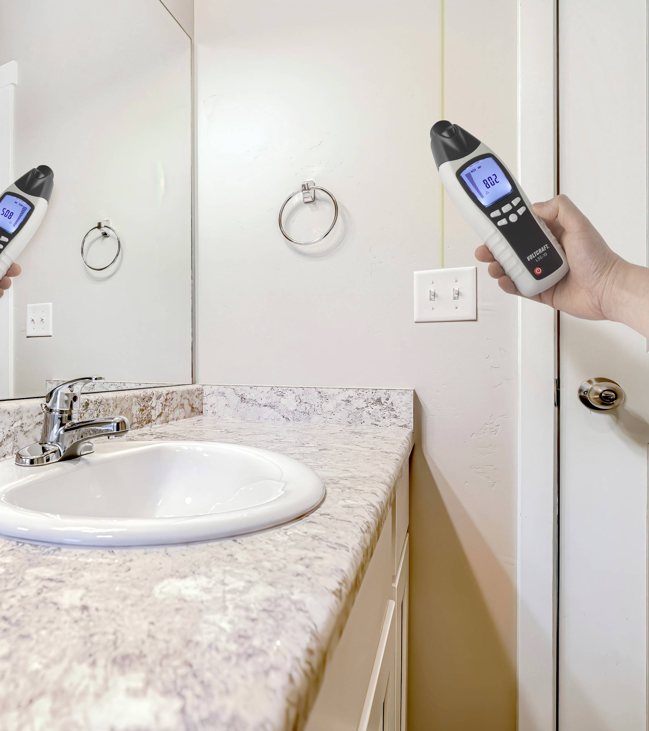 A hand holds a moisture metre over a washbasin, displaying a reading on its screen. The reflection shows the metre and the sink.