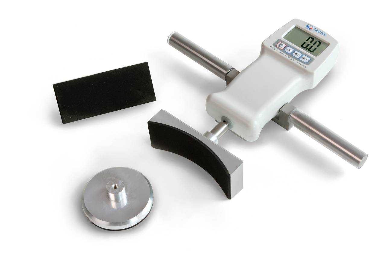 A digital measuring device with side handles, displaying a value of '0.0'. Next to it are two black and one silver metal part.