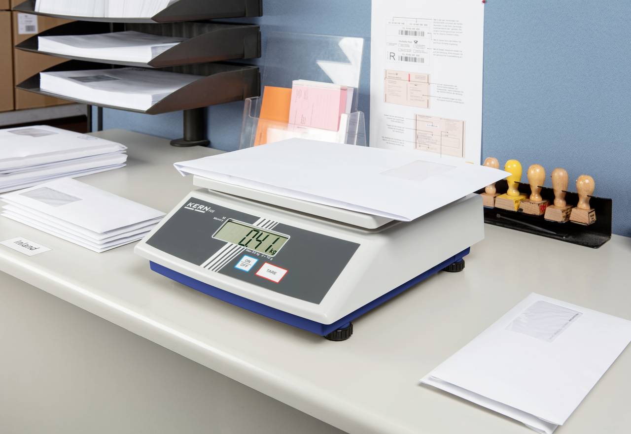A digital scales displays '0.44 kg' and is weighing a stack of letters. Postage stamps and additional envelopes are visible in the background.