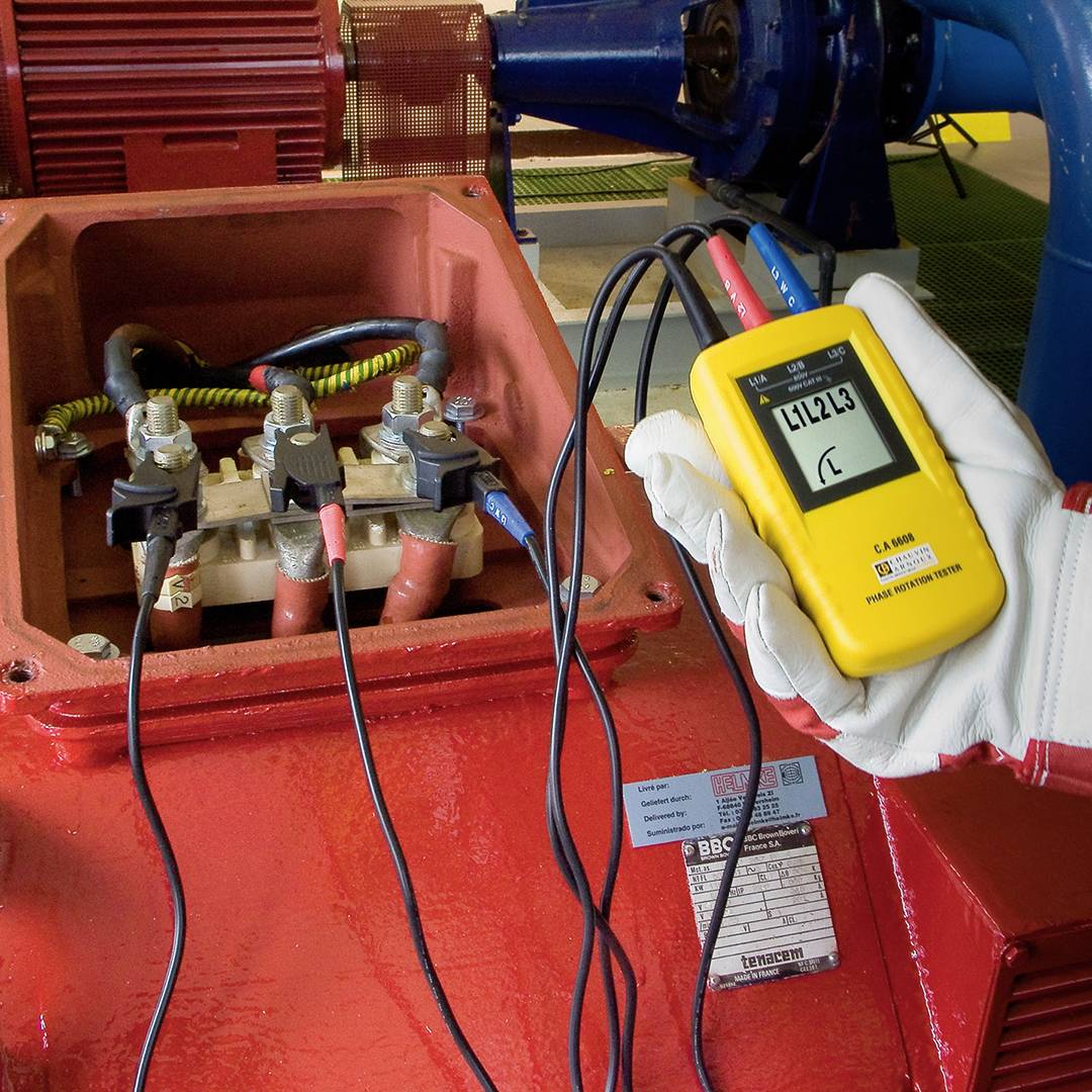 Measuring device checks the electrical current of a machine. A worker is holding the device, showing a number on the display. Red cables are connected.