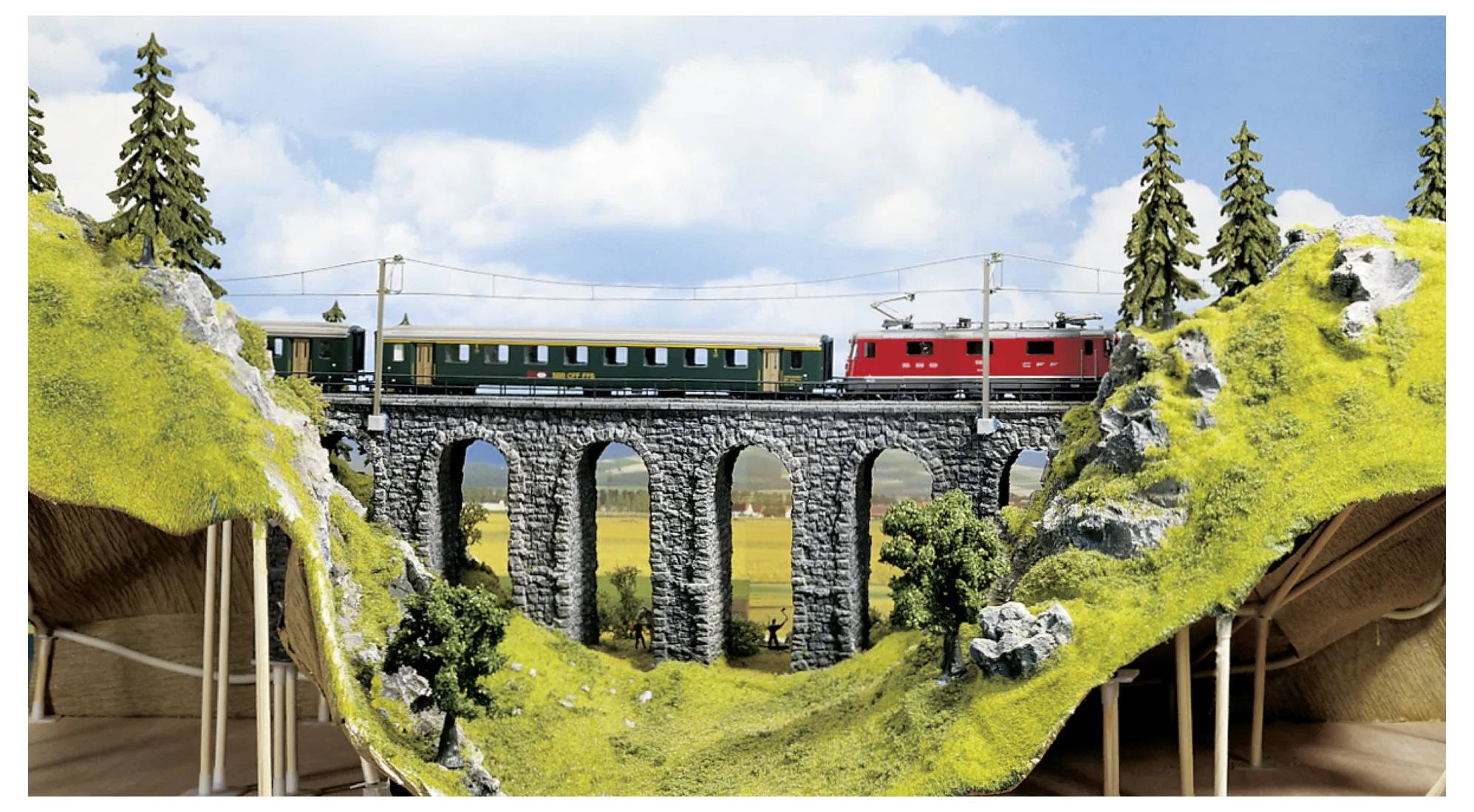A model train crossing a stone viaduct with arches, surrounded by scenic green hills and trees under a blue sky with clouds.