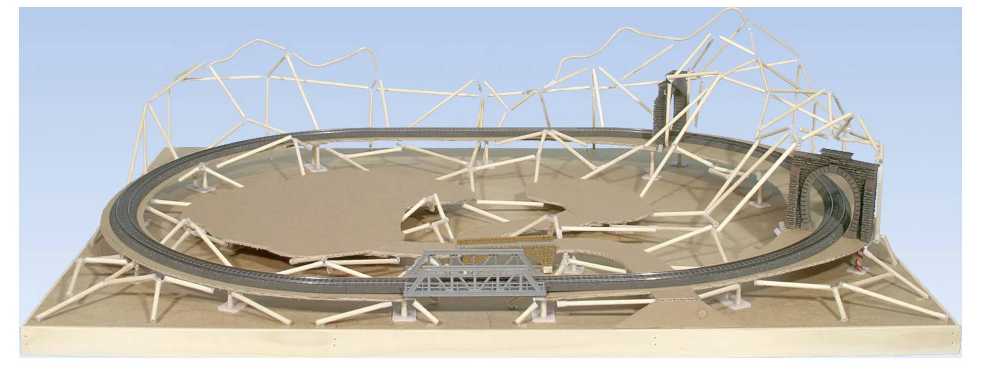 A model train layout on a plywood base, featuring a circular track with a bridge and wooden framework, simulating mountainous terrain.