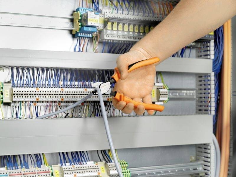 A hand holds orange-coloured pliers cutting through a grey cable inside an electrical cabinet. Laid cables and components are visible.