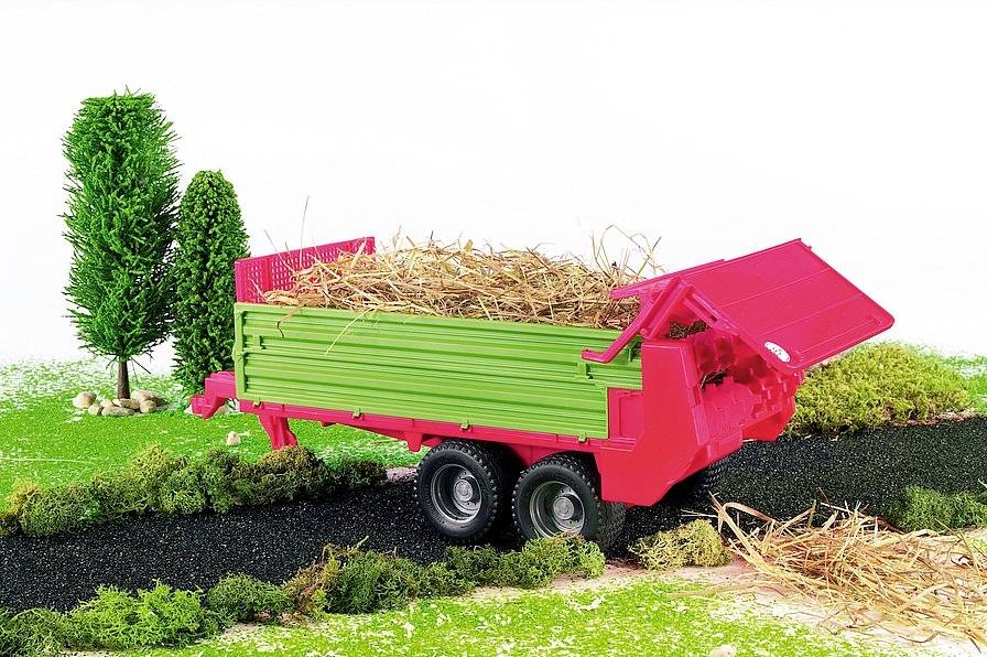 A red trailer with a green body is parked on a path, loaded with straw. Scattered bushes and trees can be seen in the background.
