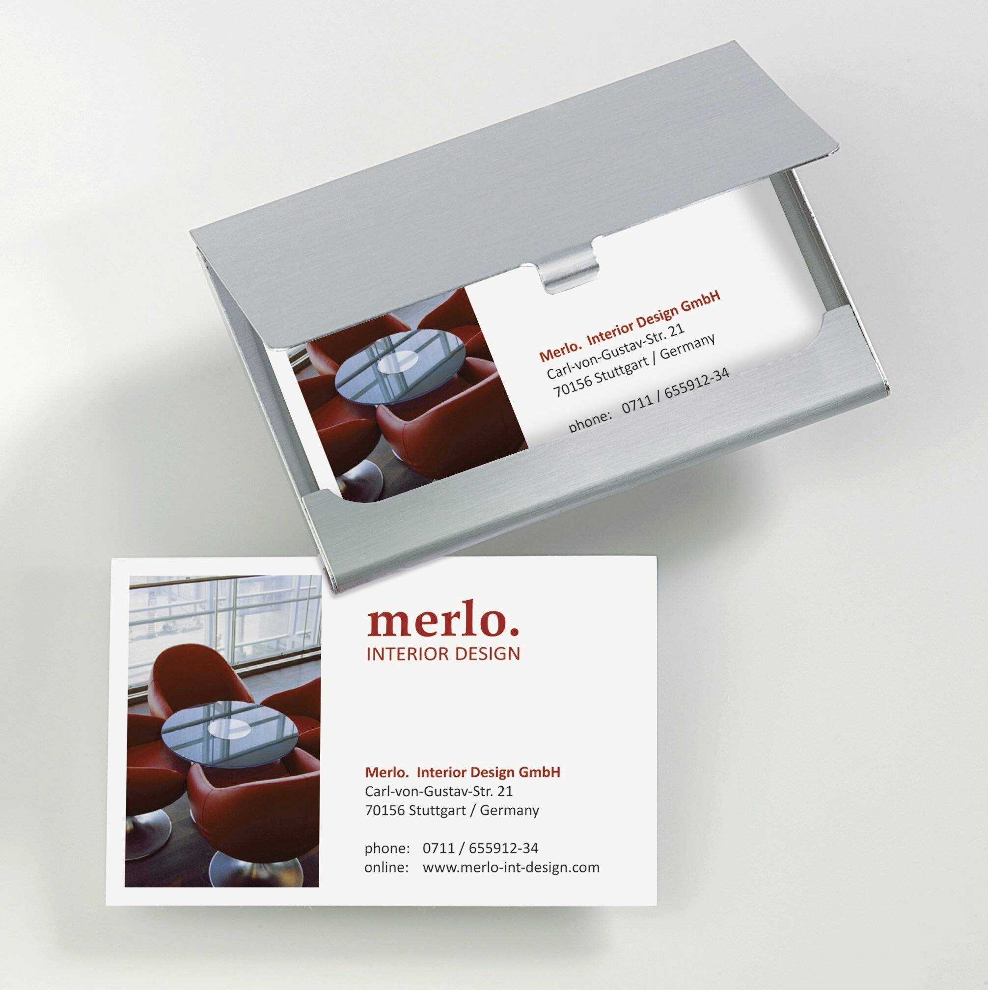 Business card for 'merlo. INTERIOR DESIGN' showing contact details and red chairs in an office setting. Card partially in a holder.