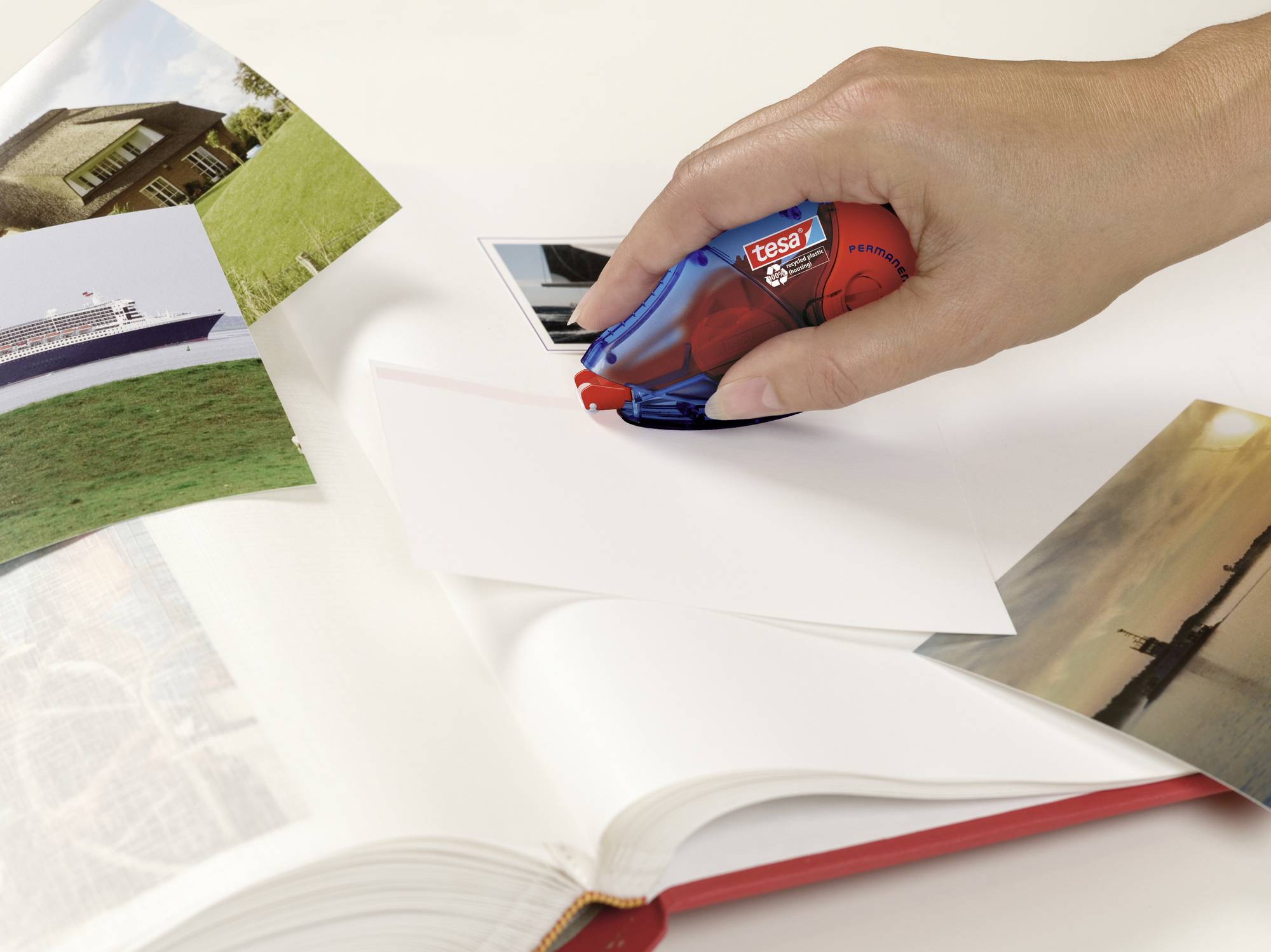 A hand is using a correction roller on paper. Photographs of a house, a ship, and a landscape are lying beside it.