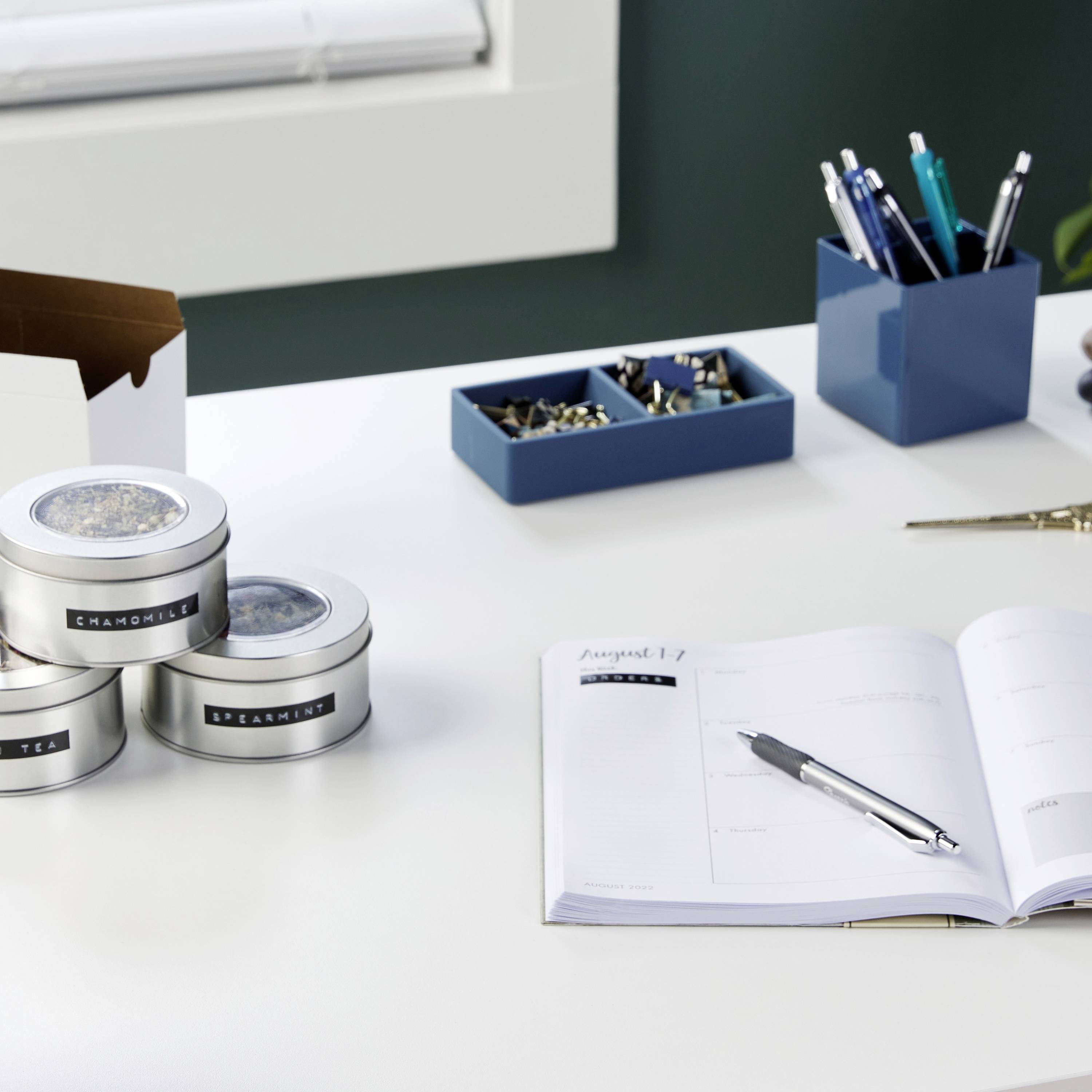 A desk with an open diary, a pen, two tins, paperclips and pens in holders.