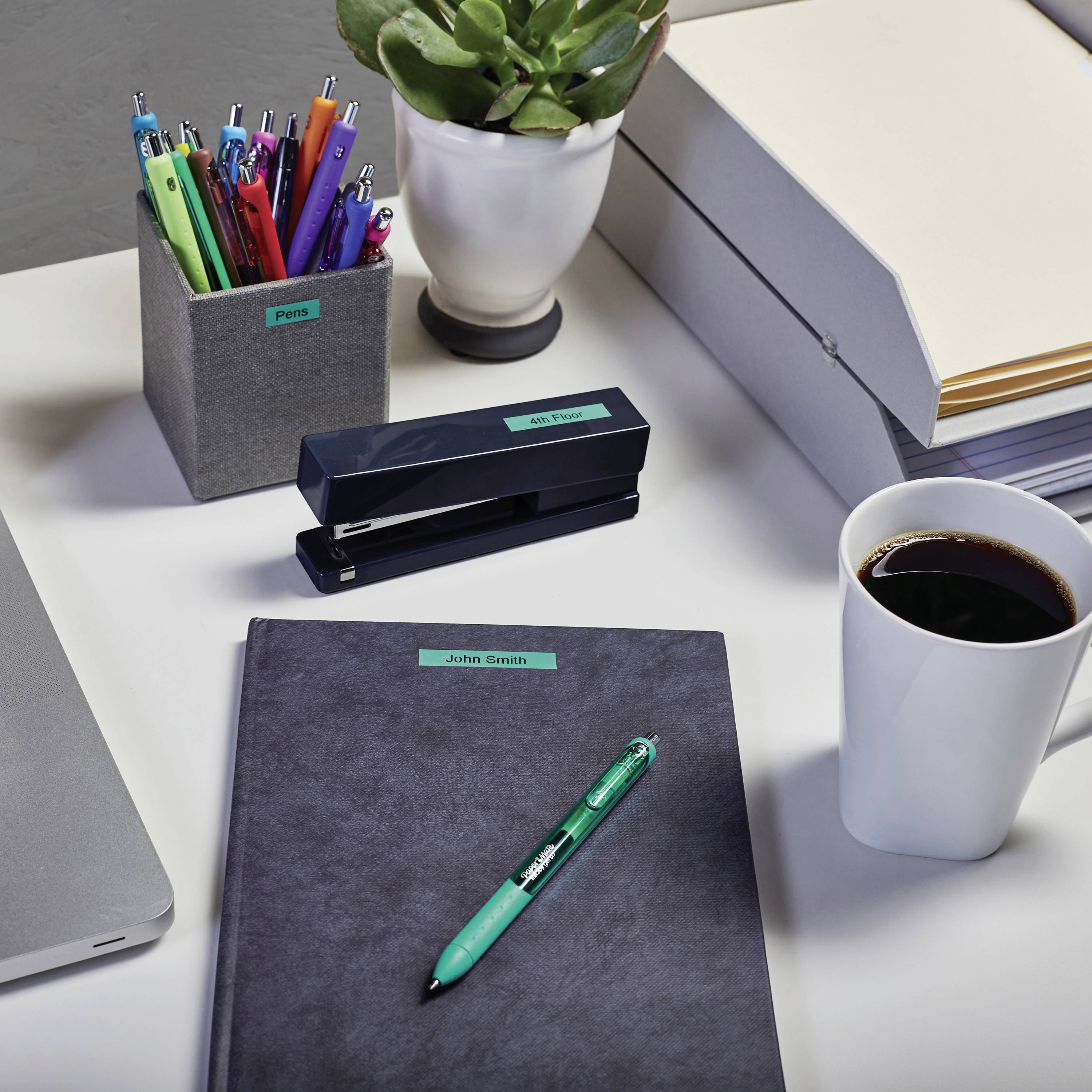A desk with colourful pens, a plant, folders, a coffee mug and a notebook with a green pen resting on top.