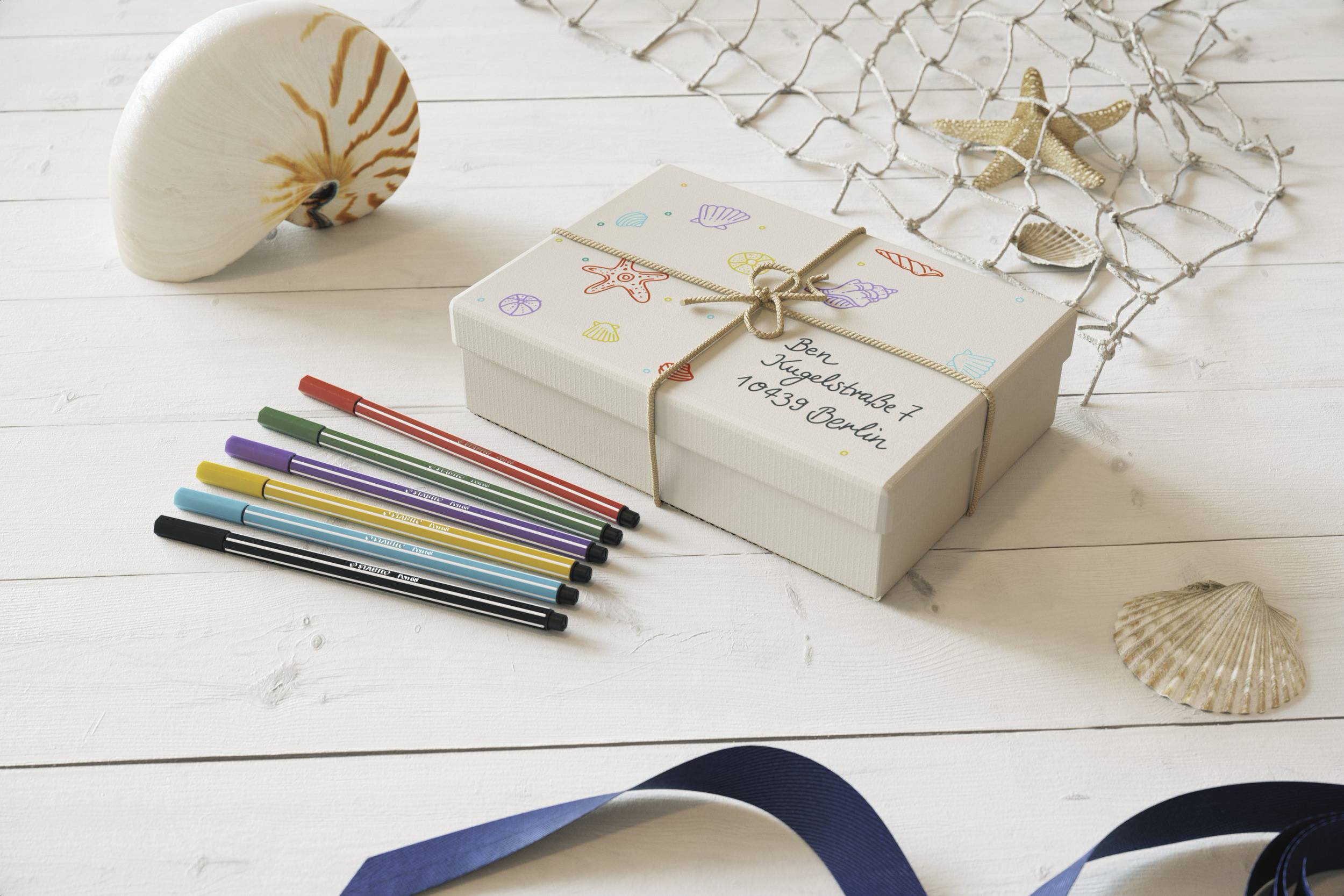 Gift box with maritime motifs and address on table, surrounded by colourful pens, shells, and a net.