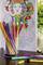 A glass filled with colourful felt-tip pens sits in front of a partially coloured colouring book with a decorative facial pattern.