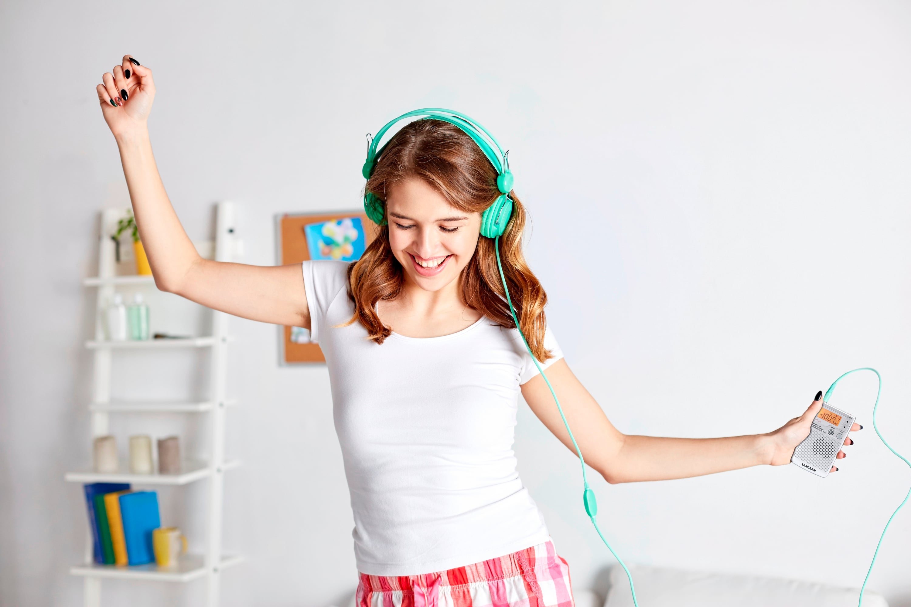 A young woman dances happily in her bedroom, wearing headphones and holding a music player in her hand.