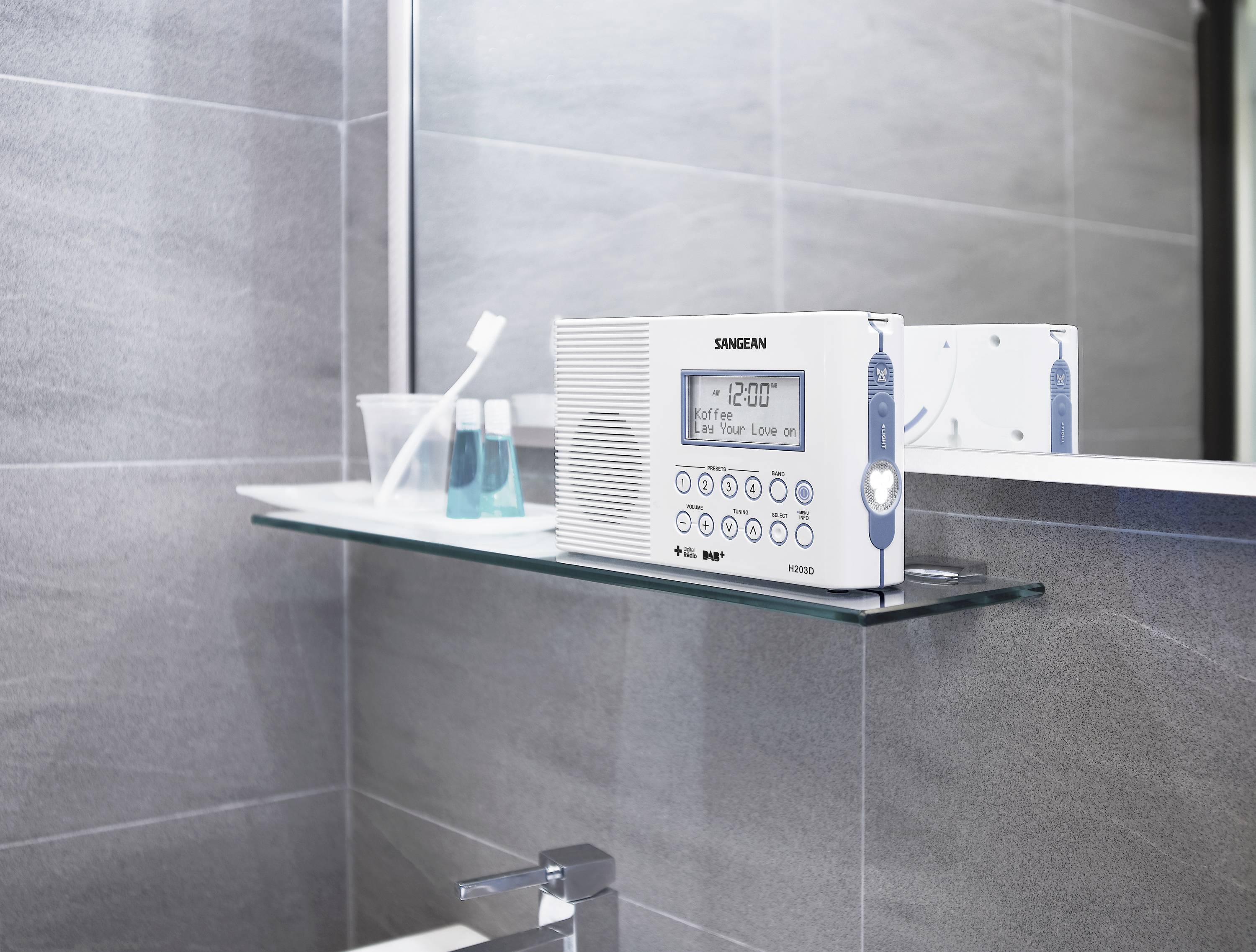 A white radio with a digital clock display shows '12:00' and sits on a glass shelf in the bathroom next to two toothbrushes and a tumbler.