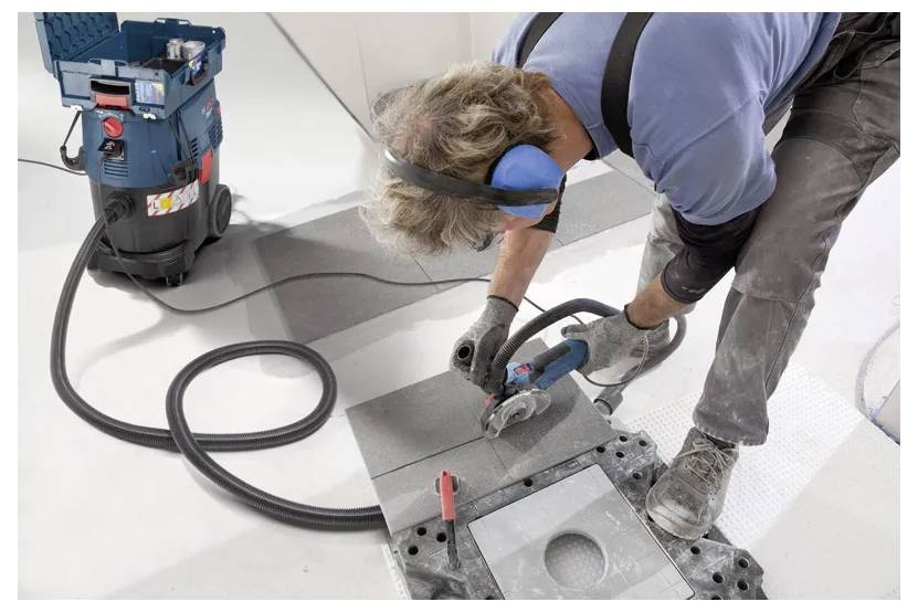 A worker wearing safety gear uses a handheld tool to cut tiles in a construction setting, with a vacuum system connected for dust control.